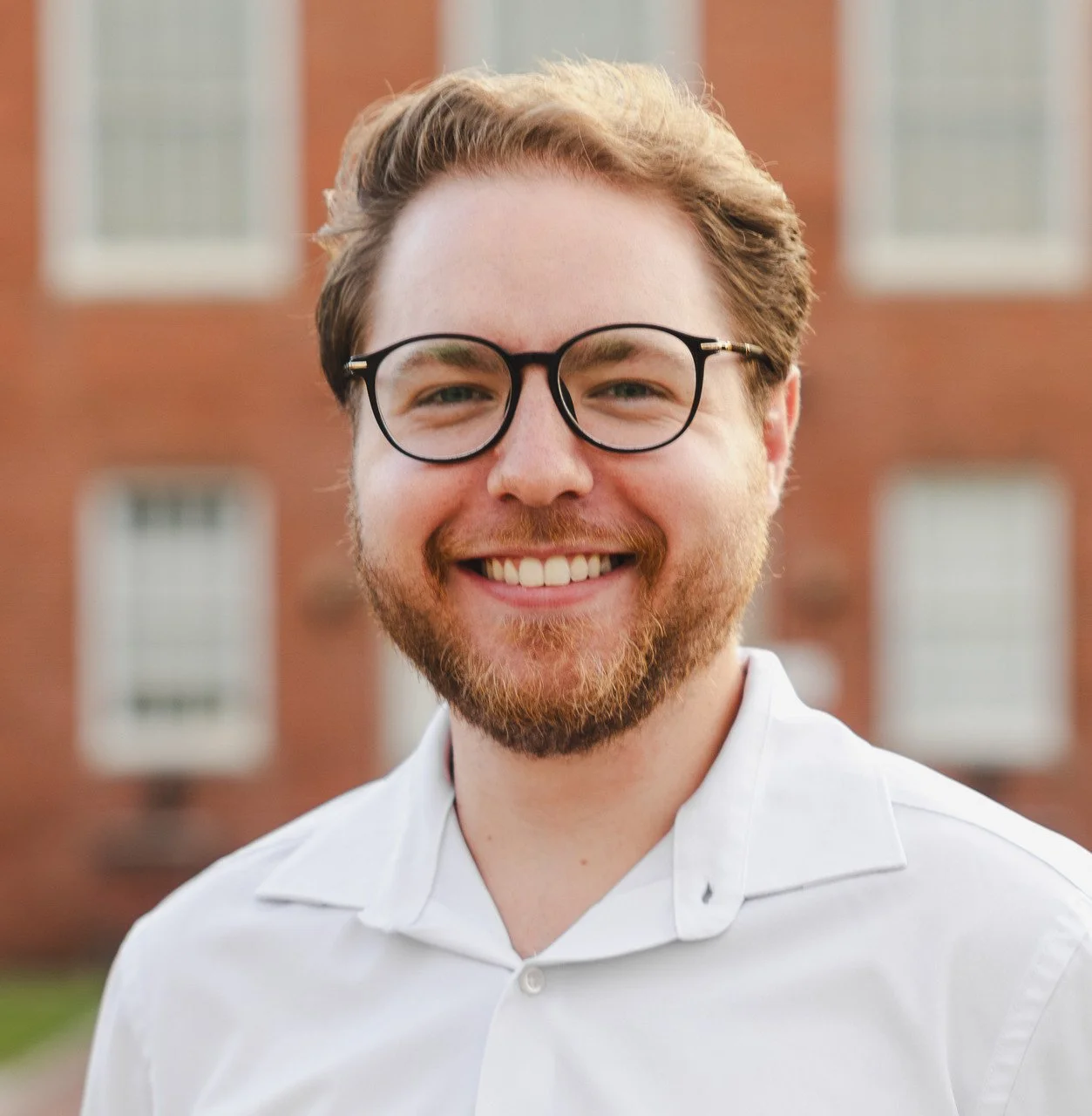 A smiling young man with glasses, red hair, and a beard, wearing a white shirt, standing outdoors in front of a red brick building.