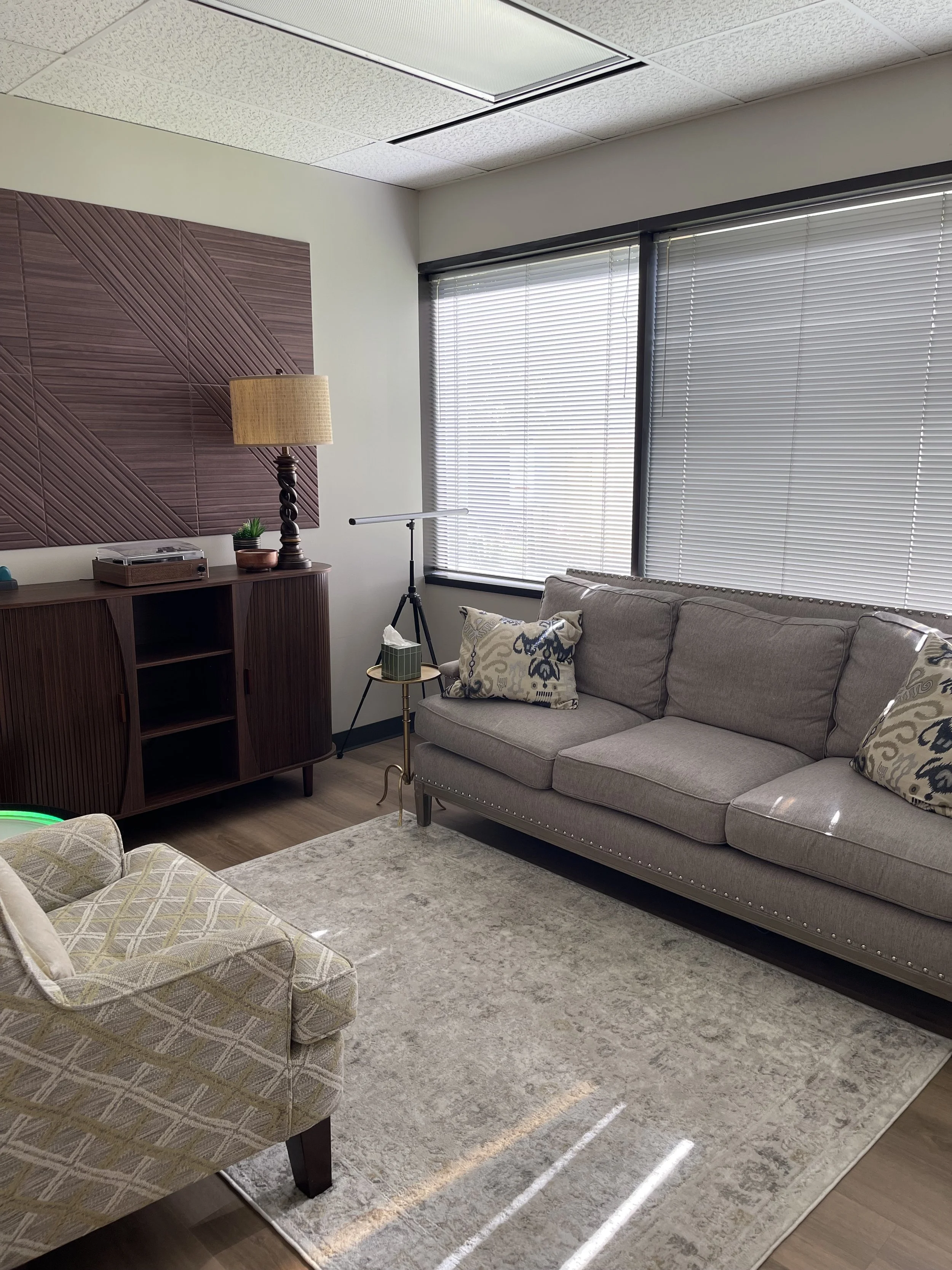 A modern living room with large windows covered by horizontal blinds, a gray sofa with decorative pillows, a wooden side table, a tall table lamp, a beige armchair, a patterned rug, and a music stand.