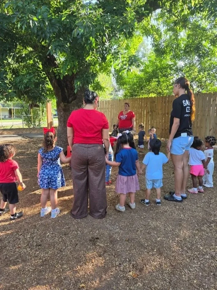 Children and adults holding hands in a circle outdoors under a large tree, with a wooden fence in the background, participating in an activity or game.