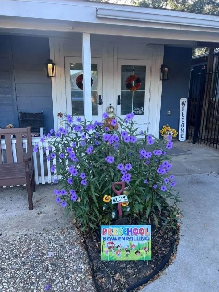 A flower bed with purple flowers and kids' Halloween decorations in front of a house. Sign in the flower bed reads "Preschool now enrolling" and a shovel with a sign saying "Hello Fall" is also in the flower bed. Wreaths are on the house's front door.