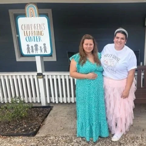 Two women standing outdoors in front of a children's learning center sign; one woman is wearing a turquoise dress, and the other is dressed in a pink tutu, white t-shirt, and tiara.