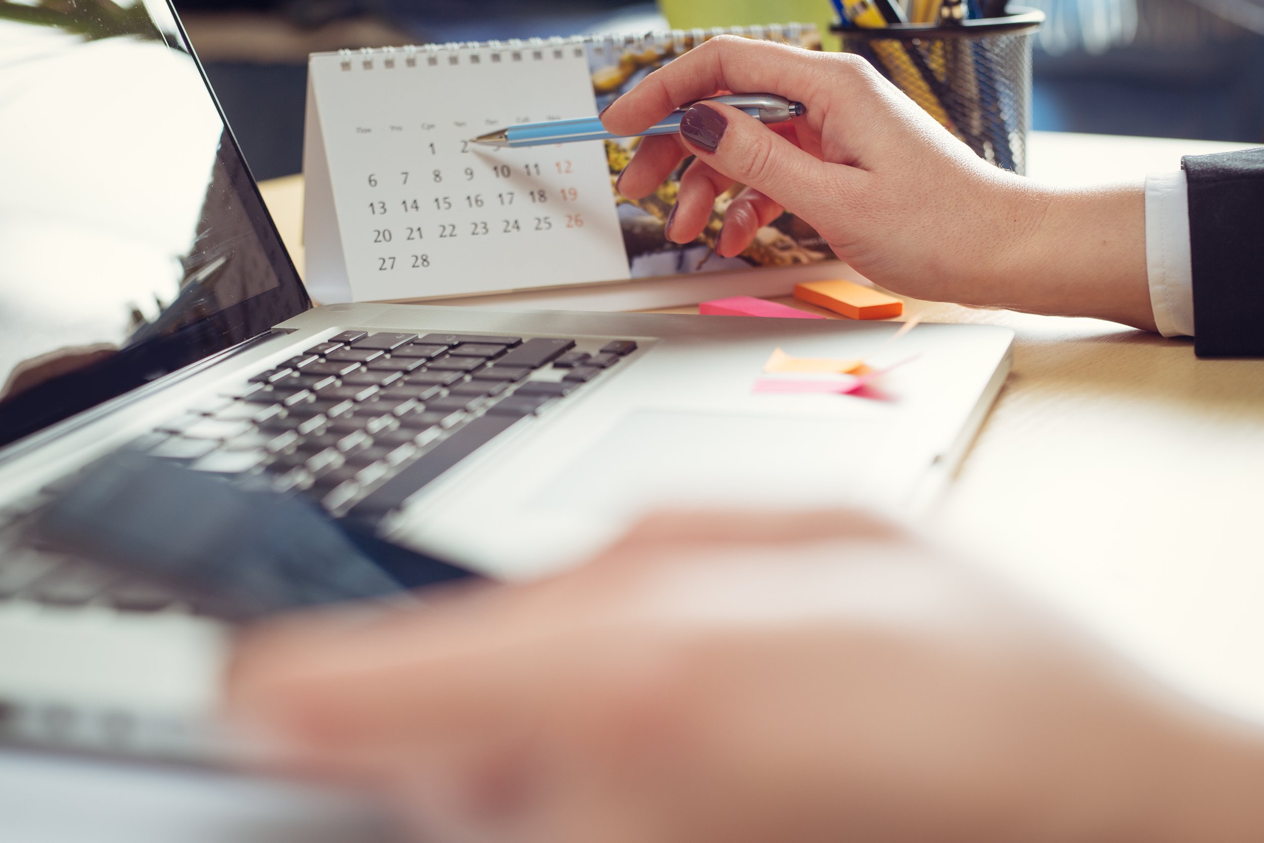 A person holding a pen and pointing at a wall calendar on a desk, with a laptop and sticky notes nearby.