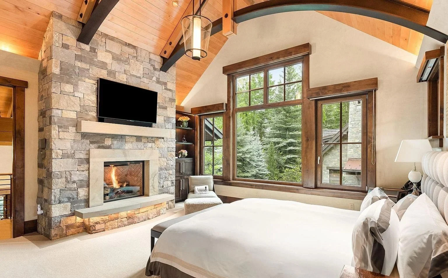 A large, cozy bedroom with vaulted ceilings featuring a stone fireplace and a spacious bed with large windows opening into a mountain forest built by John Olson, owner of Form 44 Construction, on Crystal Lake Road in Aspen, Colorado.