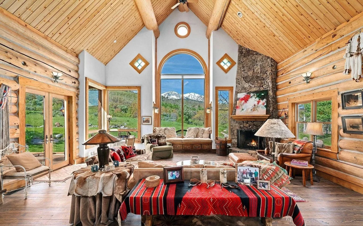 A spacious log home living room with a cozy fireplace and elegant wood floors looking out large windows on a picturesque mountain landscape built by John Olson, owner of Form 44 Construction, on Old Herron Road in Basalt, Colorado.