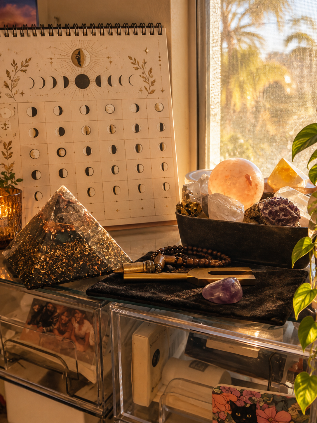 A desk displaying various crystals, a tarot card measuring board, and a pyramid-shaped resin art piece with gold flakes, with a sun and moon calendar on the wall and a window showing trees outside.