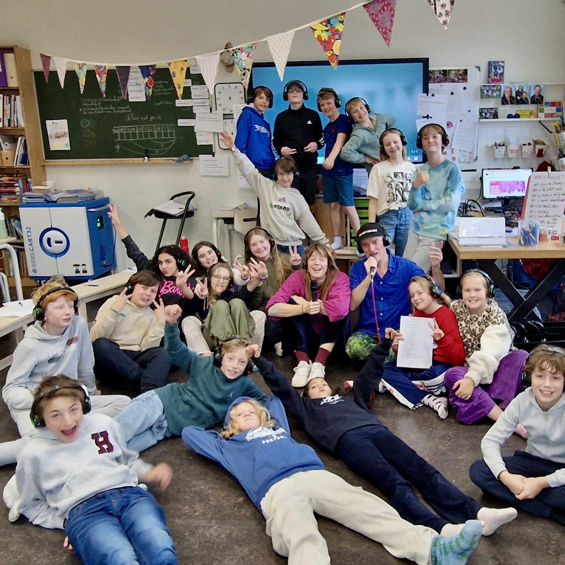 Een klaslokaal met kinderen die lachen en poseerden voor de camera. Sommige dragen koptelefoons. Er hangen kleurrijke vlaggetjes aan het plafond, en er is een bord met schoolmateriaal en een groot scherm op de muur.