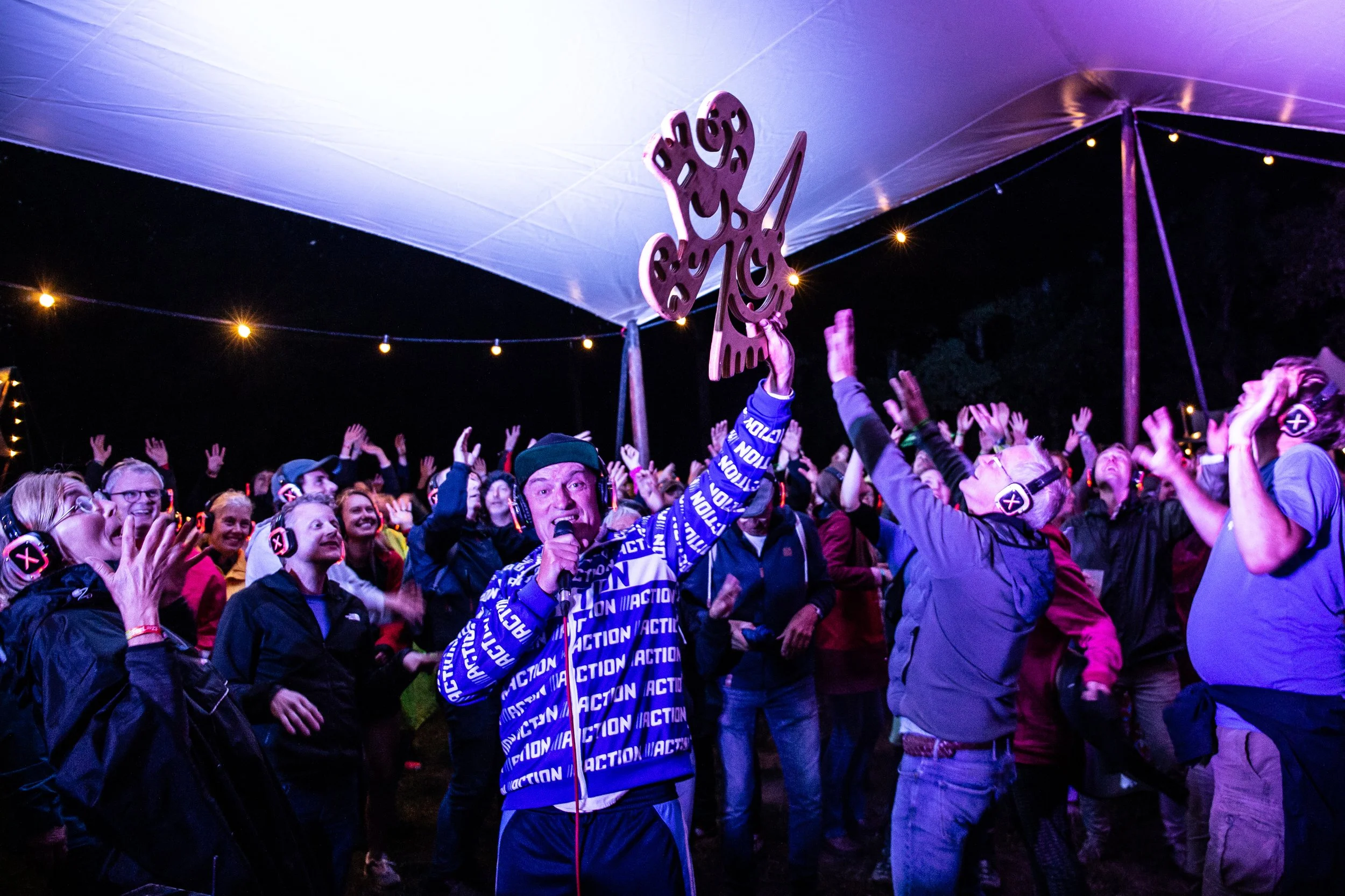 Grote groep mensen bij een nachtelijk evenement onder een tent, met de aandacht op een man die een houten vlinder vasthoudt en spreekt in een microfoon.
