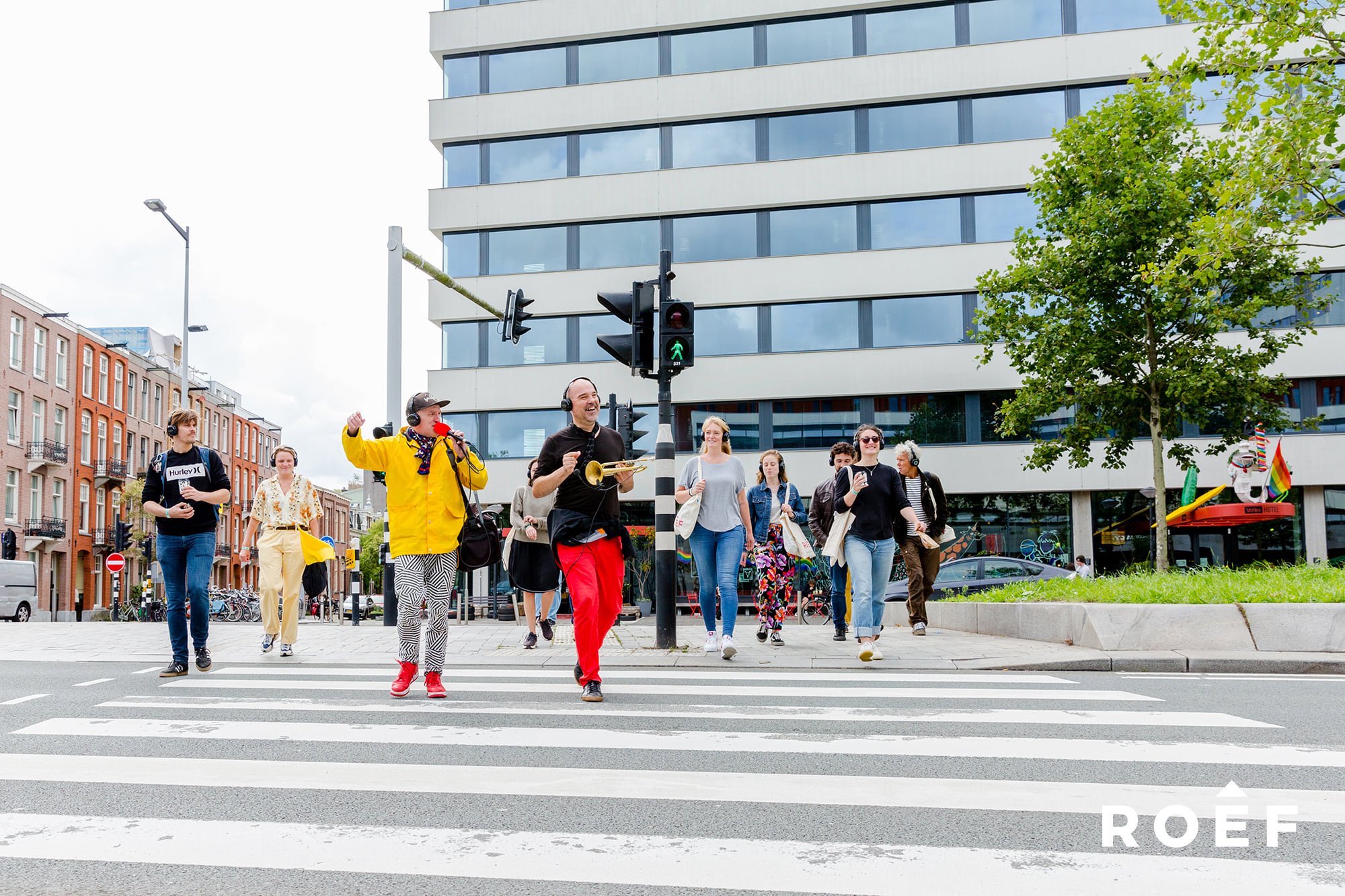 Mensen lopen over zebrapad in de stad, een muzikant speelt trompet, mensen dragen koptelefoons en dragen kleurrijke kleren, verkeerslichten en moderne gebouwen op de achtergrond.