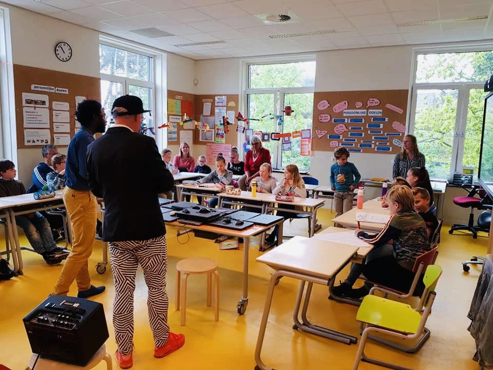 Een klaslokaal met leerlingen en leraren, mensen staan en zitten, decoraties aan de muur, grote ramen met uitzicht op bomen, leerlingen lijken deel te nemen aan een interactieve sessie.