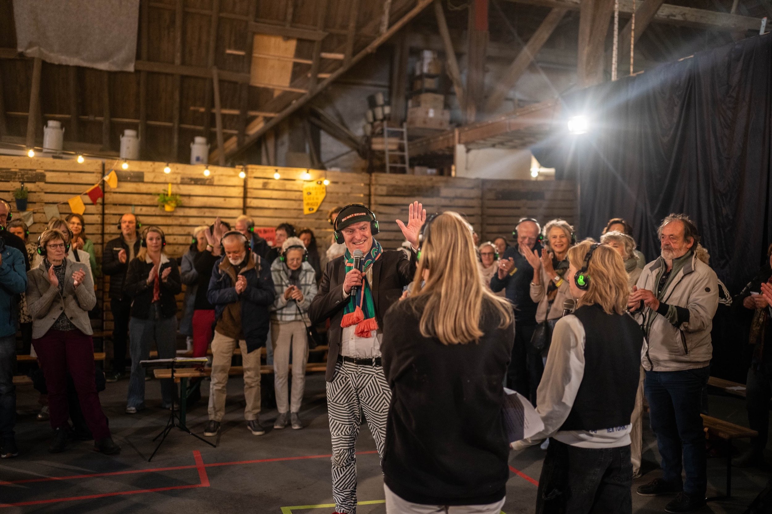 Binnen een grote loods een groep mensen die naar een man met een microfoon kijken. De mensen dragen hoofdlampjes of koptelefoons en applaudisseren, de sfeer is blij en feestelijk.