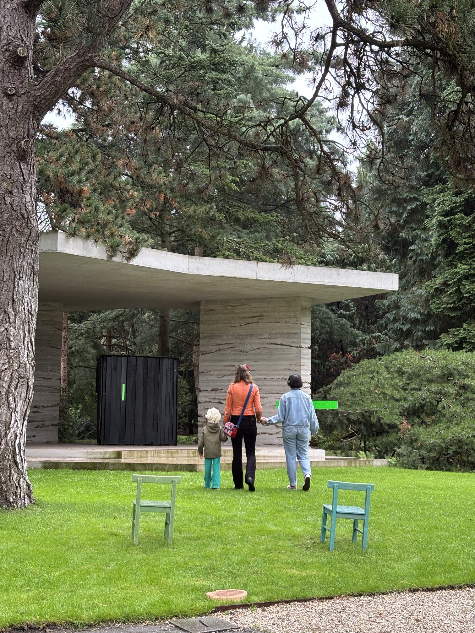 Twee volwassenen en twee kinderen lopen hand in hand in een park met groene gras, bomen en een moderne architectonische structuur op de achtergrond.
