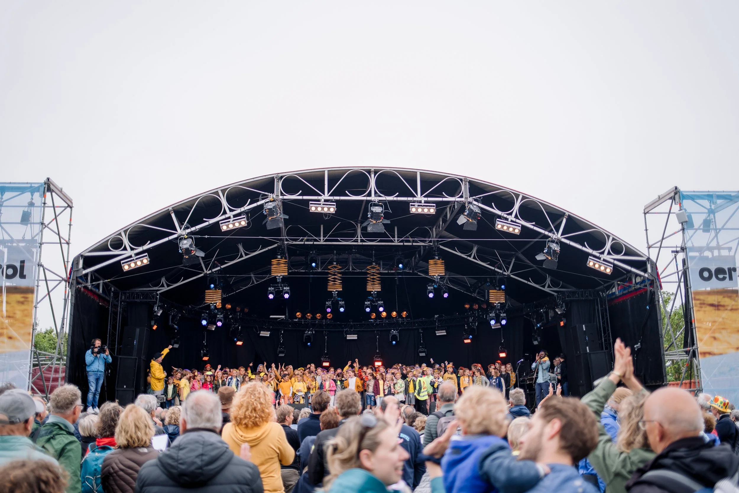 Grote menigte mensen bij een openluchtconcert met een groot podium en band, onder een witte hemel.