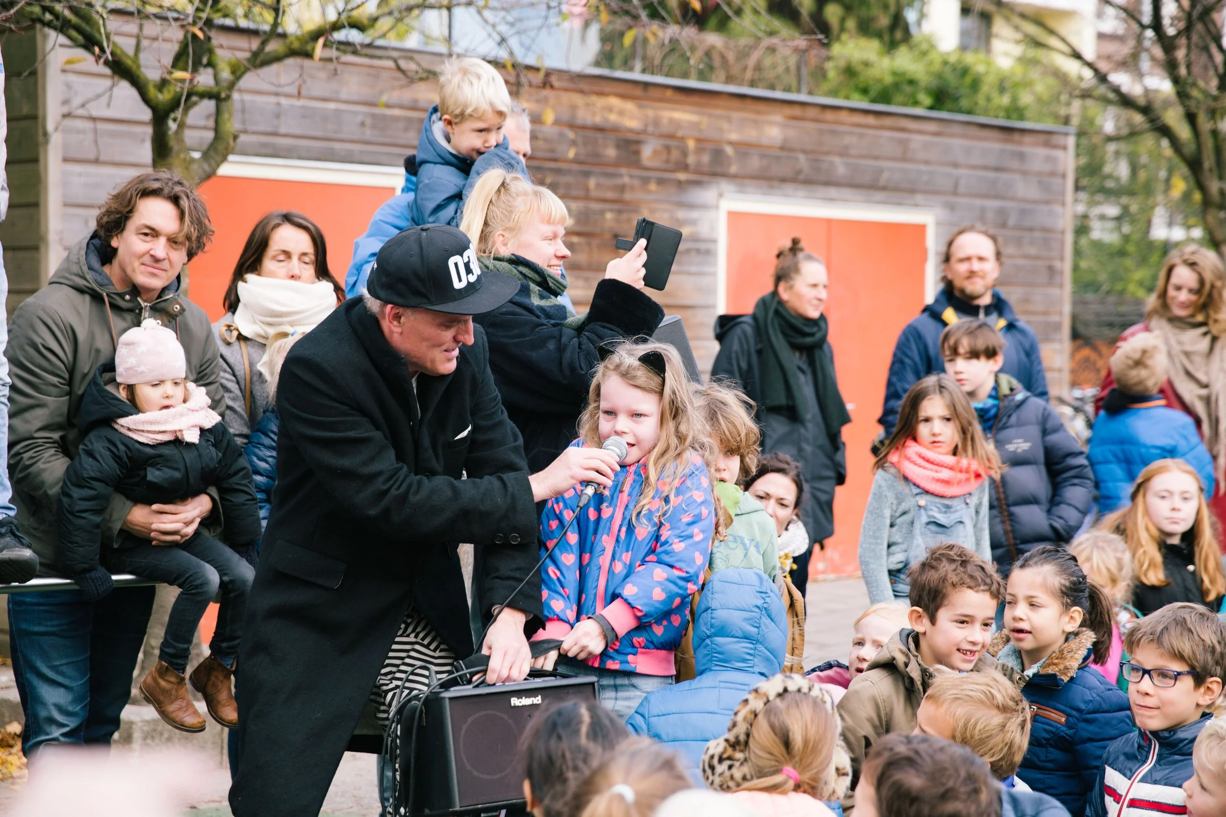 Een groep volwassenen en kinderen samen in een openluchtreliëf, met veel mensen die luisteren en meedoen, onder een boom en een houten gebouw. Kinderen spreken en zingen, volwassenen kijken en luisteren naar een optreden.