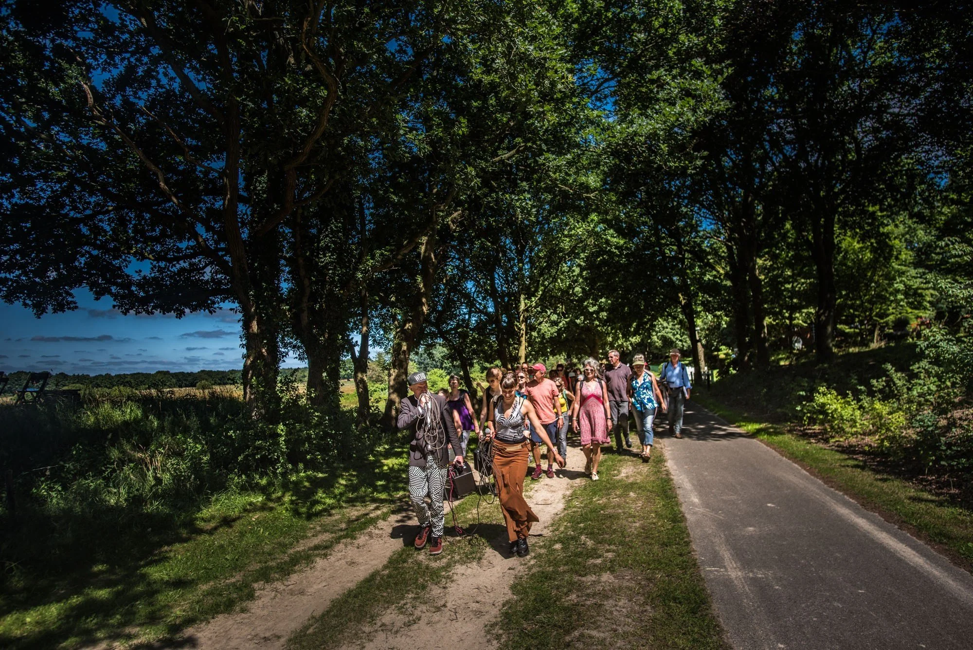 Groep mensen wandelt langs een pad onder een bosrijke boomgaard op een zonnige dag.