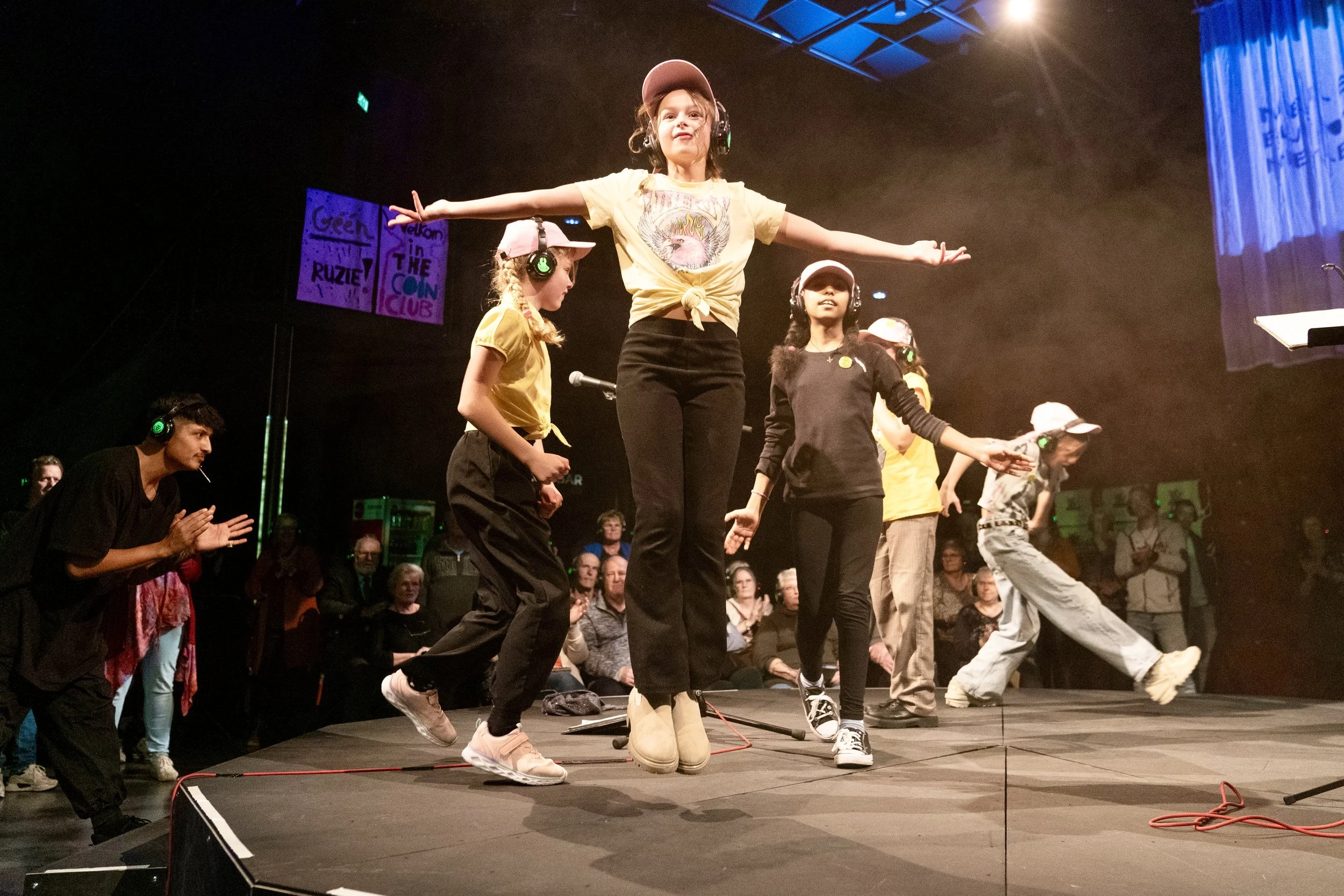 Kinderen en volwassenen op een podium, dansend en plezier hebbend tijdens een optreden. Mensen kijken toe en applaudisseren op de achtergrond.