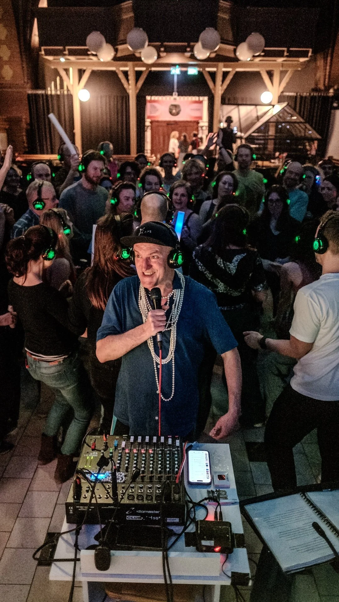Een DJ staat achter een mengpaneel met koptelefoons en een microfoon in een levendige danshoek, omringd door feestvierders met koptelefoons.
