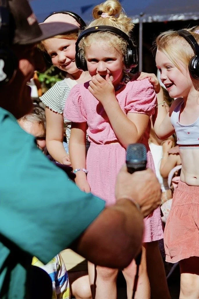 Groep meisjes met koptelefoons luisteren naar een interviewer met een microfoon tijdens een buitenactiviteit.
