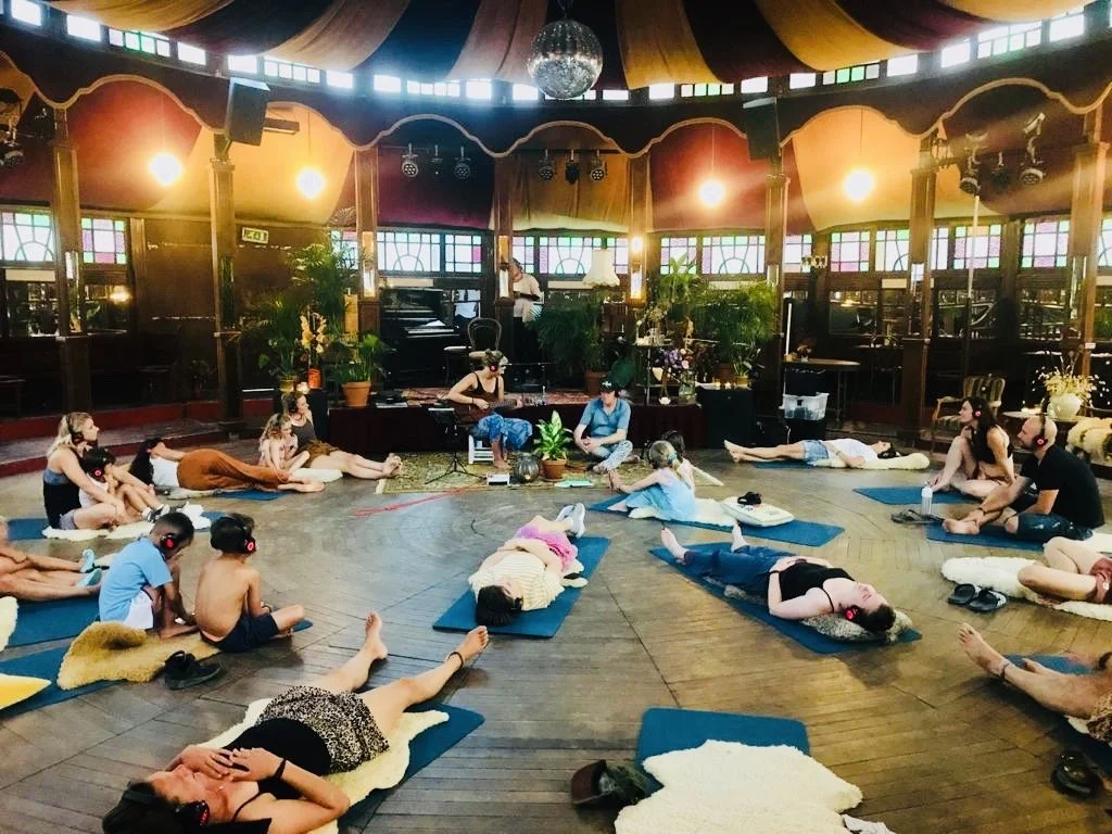 People, inclusief kinderen, doen yoga of meditatie op matten in een ruimte met kleurrijke ramen en decor. Ze dragen koptelefoons en liggen of zitten in verschillende rustposities.