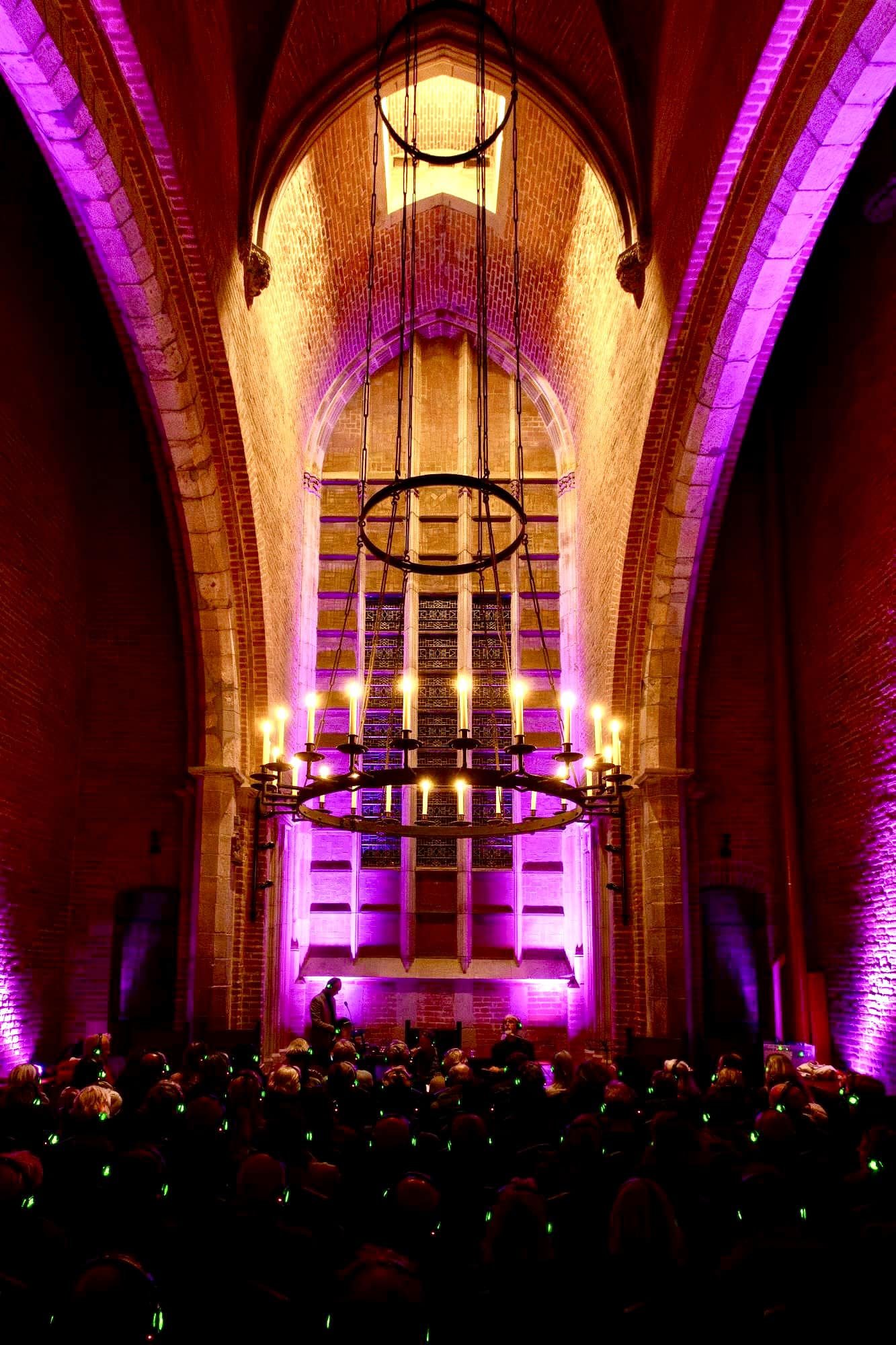 Een kerkinterieur met hoge, gebrandschilderde ramen en een grote, ronde kroonluchter met kaarslichtekeningen. De muren worden verlicht in roze en paarse tinten. Er zit een menigte mensen met oortjes op, die lijkt te luisteren naar een lezing of presentatie.