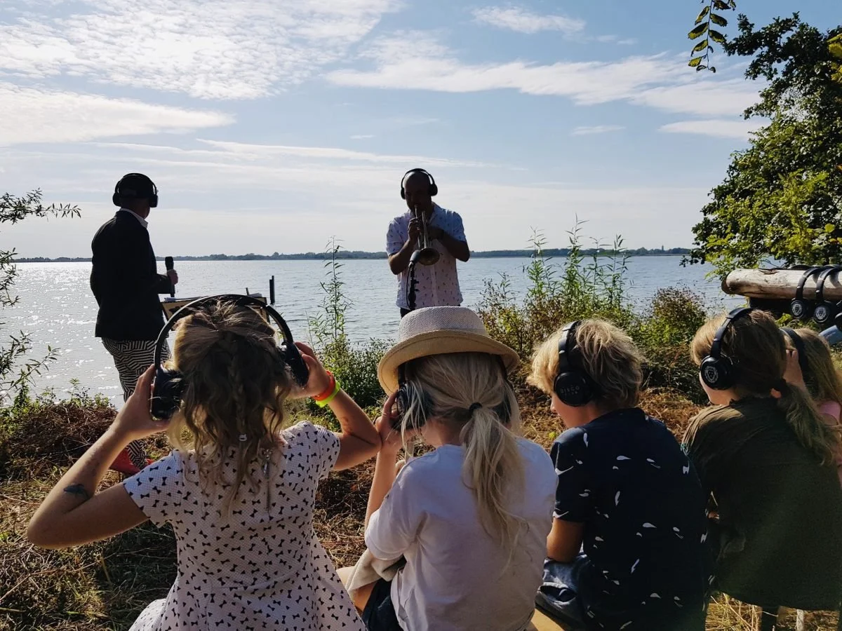 Kinderen luisterend naar een optreden bij een meer, iedereen draagt koptelefoons, twee mannen voerend de voorstelling, het is een zonnige dag met lichte bewolking.