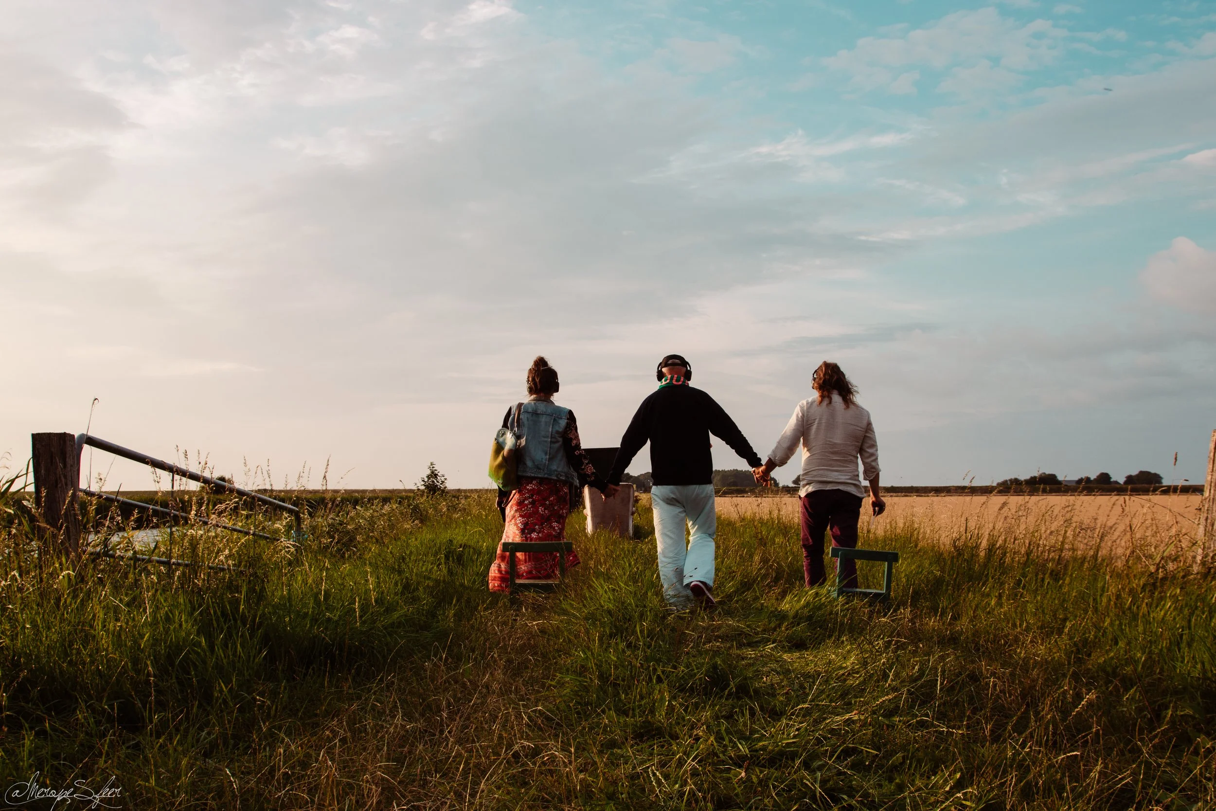 Drie mensen wandelen hand in hand door een veld onder een bewolkte hemel, met een hek en veel gras op de voorgrond.