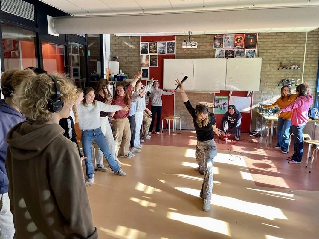 Groep jongeren in een klas of aula, met een meisje dat danst terwijl anderen kijken en meezingen, in een ruimte met posters op de muur en grote ramen.