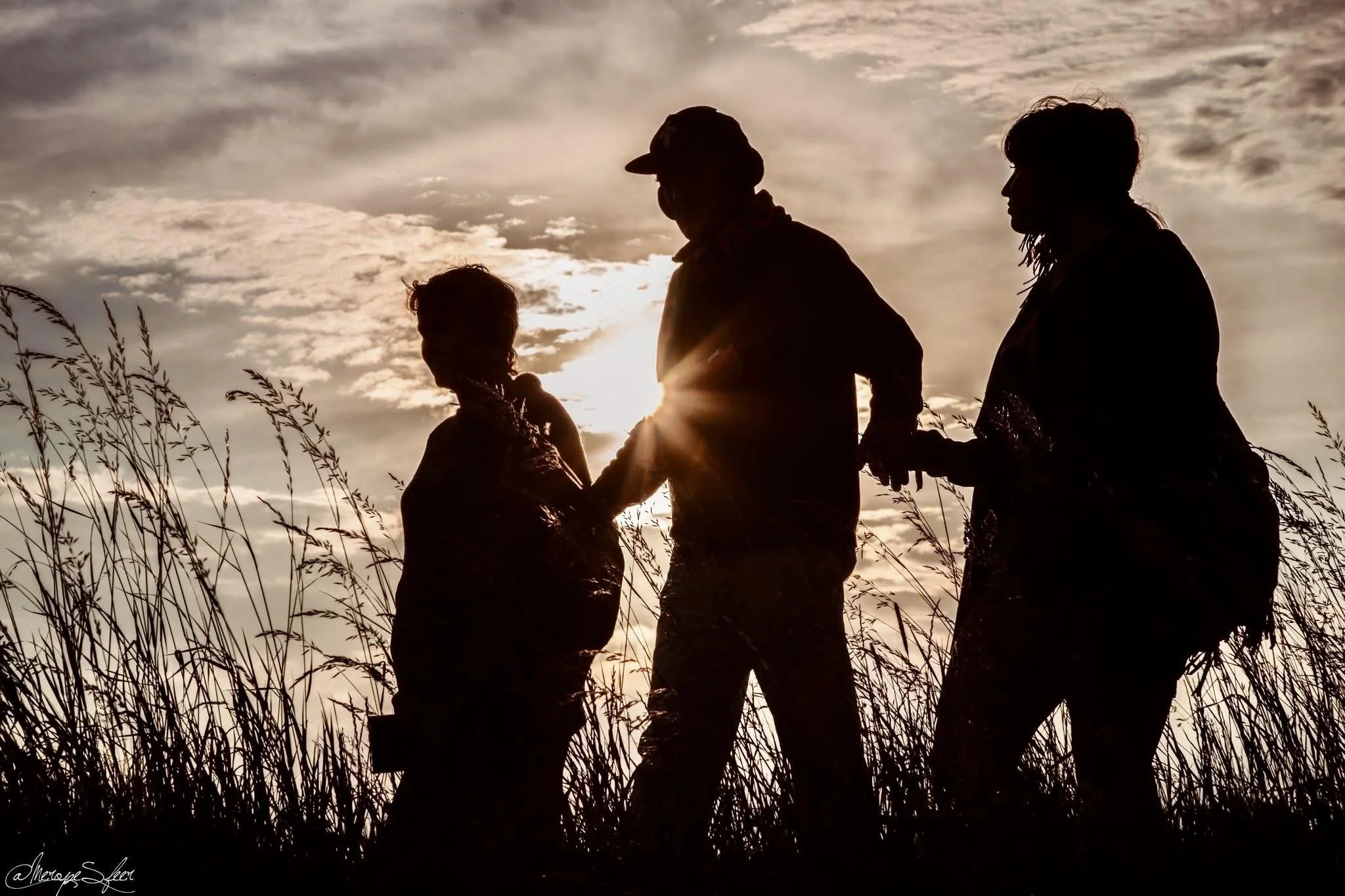 Silhouet van vier mensen die hand in hand staan in een veld bij zonsondergang, met een bewolkte hemel.