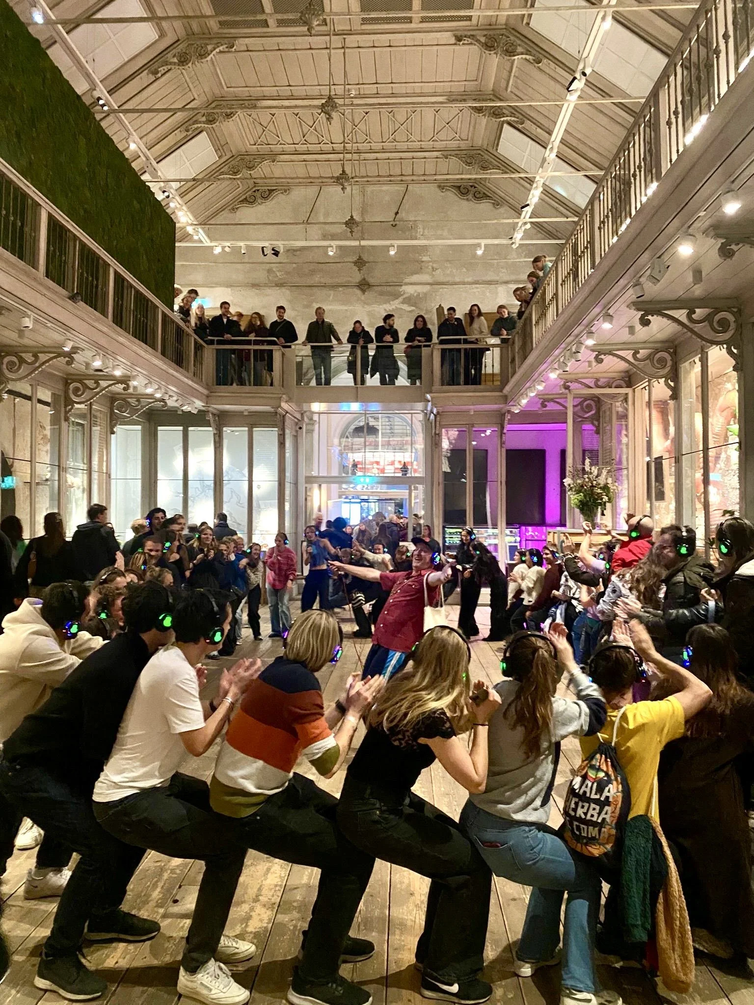 Groep mensen die een polonaise dansen in een grote, gezellig ingerichte zaal met twee verdiepingen.