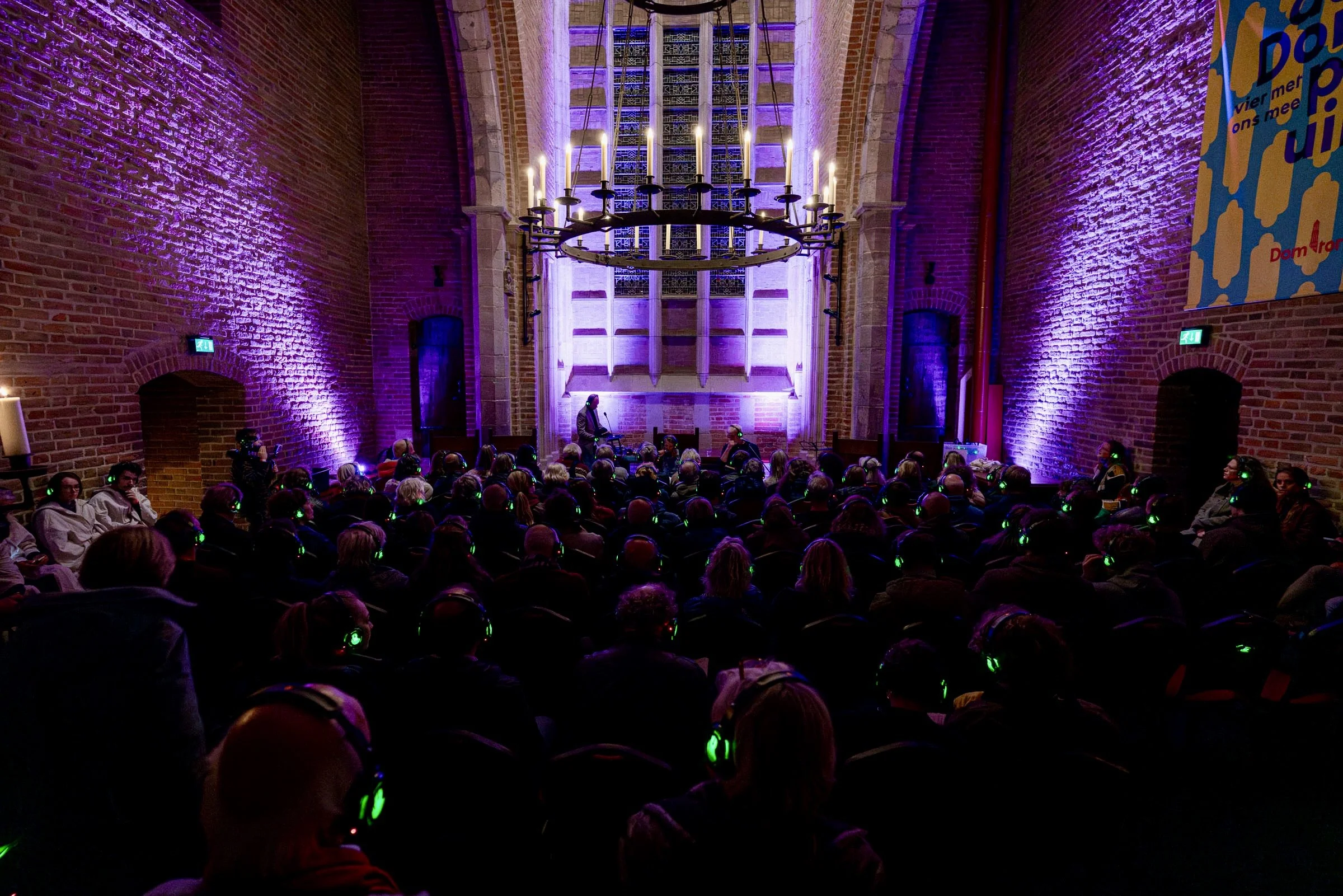 Binnen in een kerk met veel publiek, een grote ronde kroonluchter met kaarsen hangt aan het plafond, blauwe en paarse verlichting verlicht de bakstenen muren.