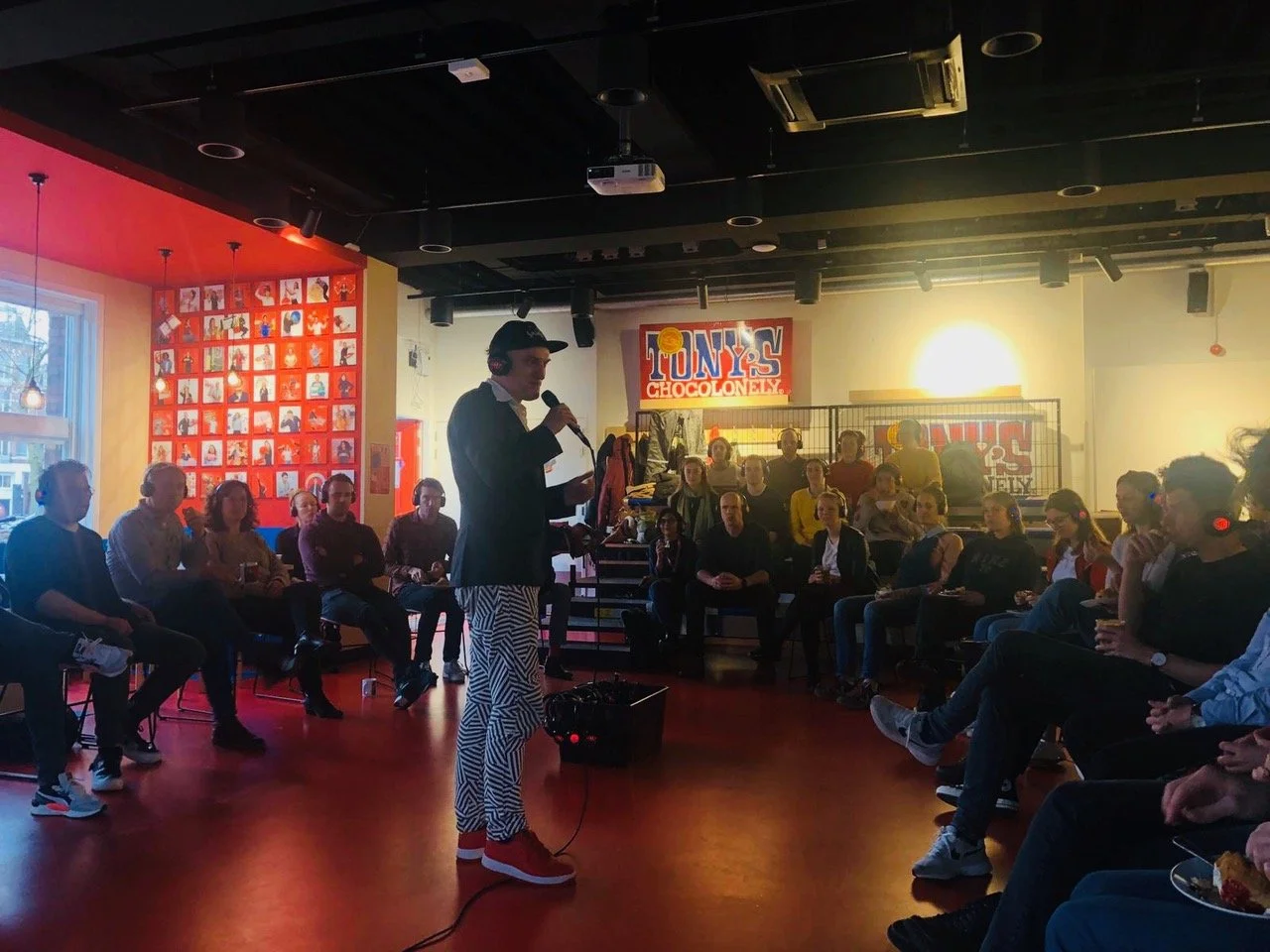 Een man in zwaren enbaan met een microfoon spreekt voor een groep mensen in een zaal. Op de achtergrond is een bord met ‘TONY’S CHOCOLONELY’ en een muur met foto's.