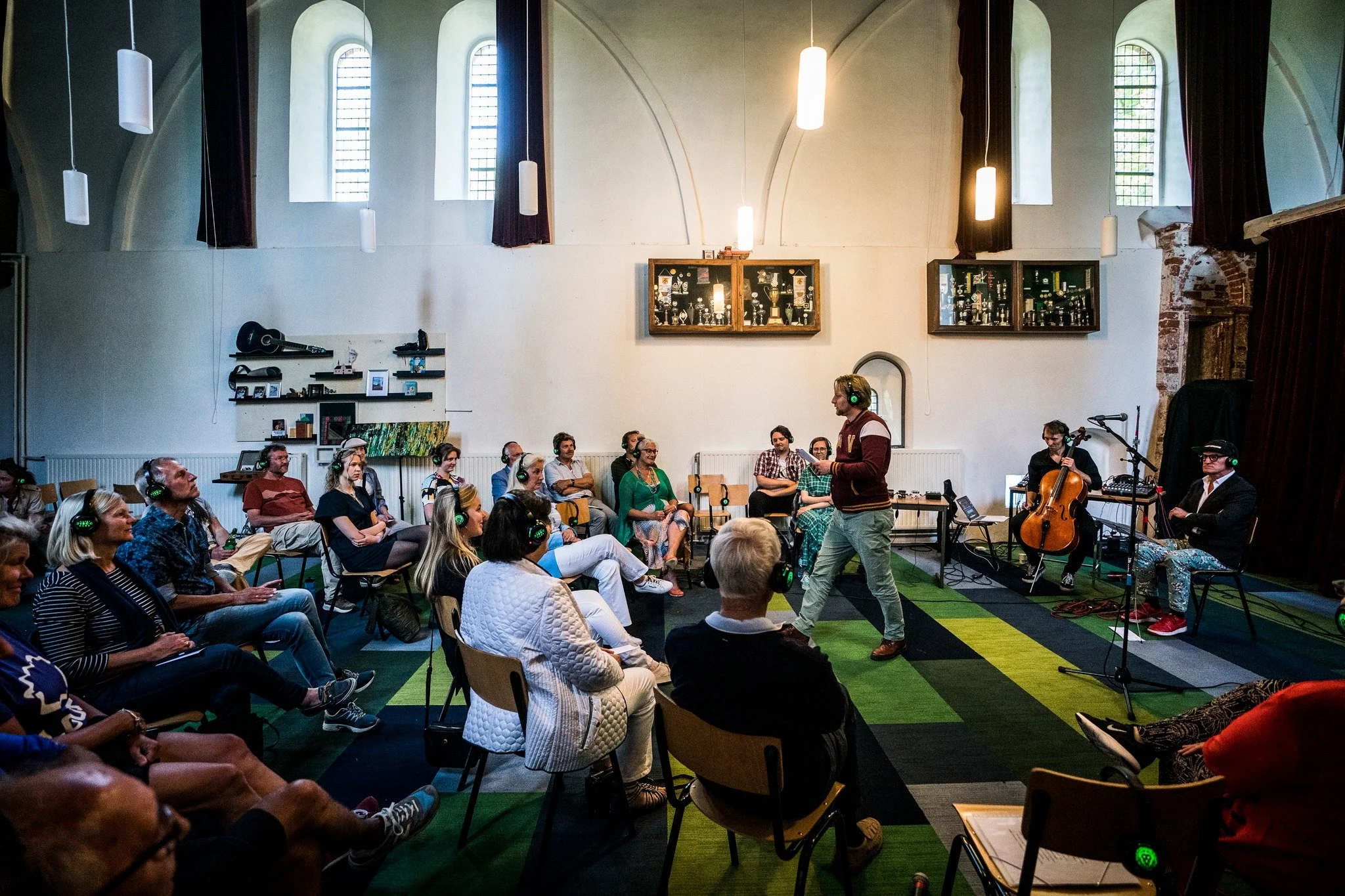 Een groepsbijeenkomst in een kerk met mensen die luisteren naar een spreker. Er zijn muzikanten met instrumenten en koptelefoons, en het publiek zit in stoelen of op de grond.