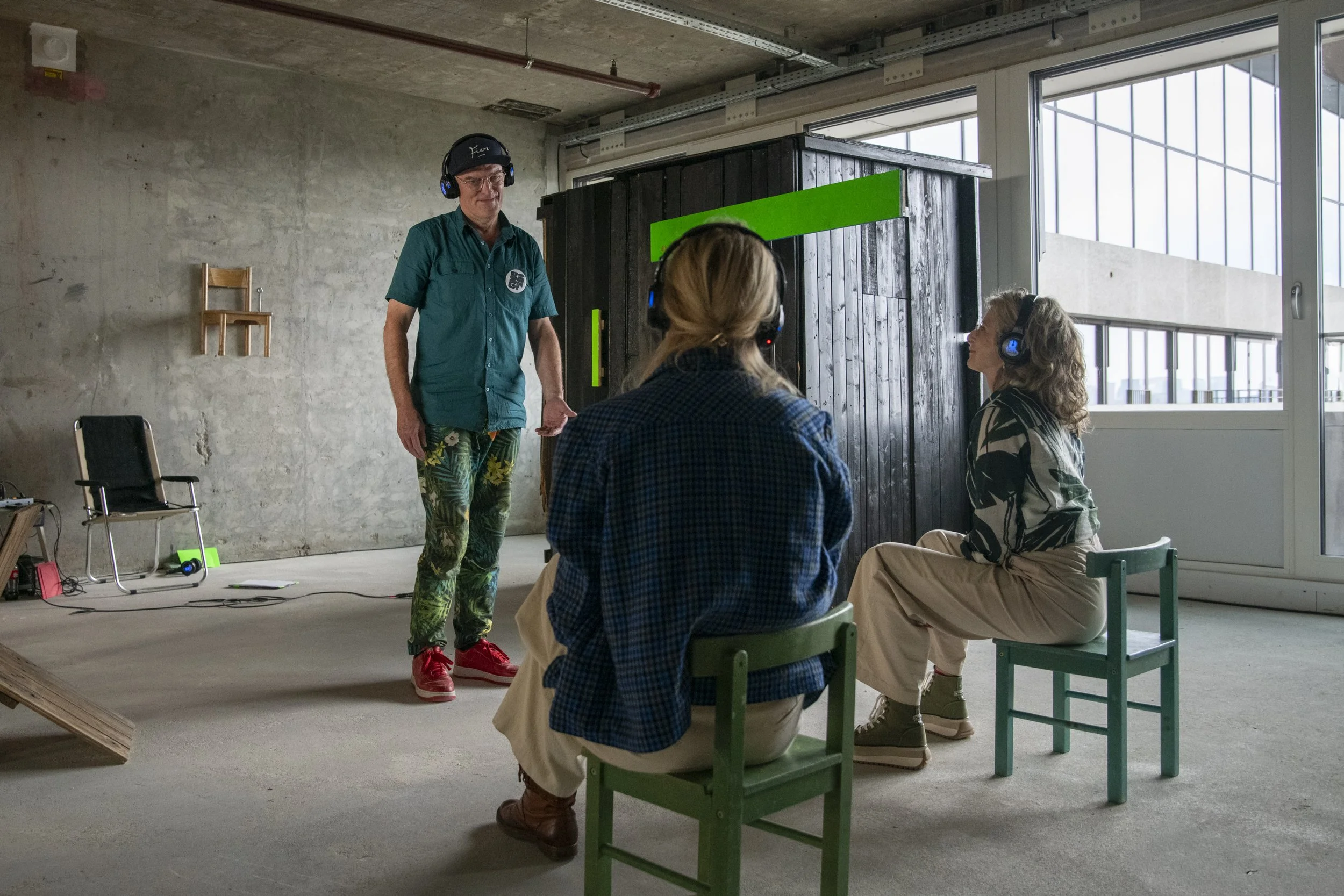 Drie mensen met hoofdtelefoons in een kamer met een betonnen muur, één persoon staat terwijl twee anderen zitten op stoelen. Een persoon steekt de hand uit tijdens een gesprek of presentatie.