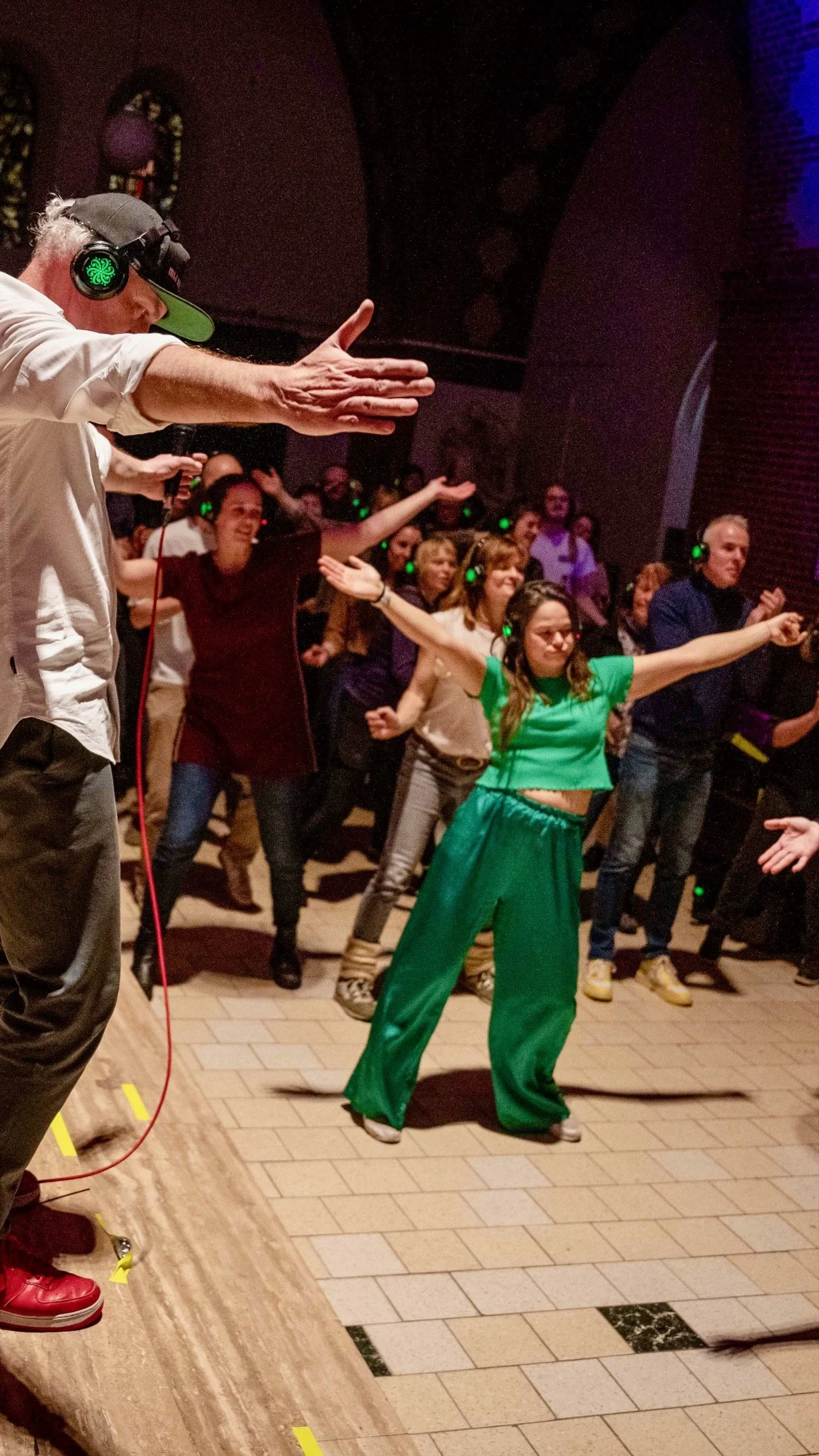 Een groep mensen danst en geniet op een dansvloer, met een vrouw in het groen die in het middelpunt staat. Mensen dragen koptelefoons, waarschijnlijk tijdens een silent disco.
