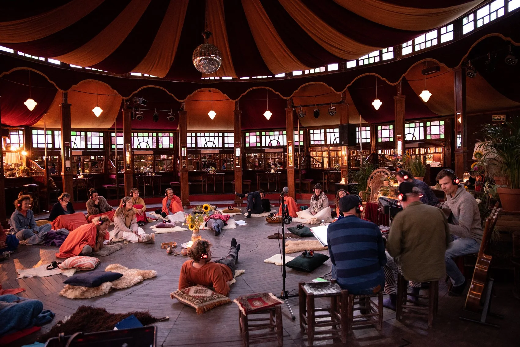 Mens en vrouwen zitten op de grond tijdens een yogales of meditatie sessie in een grote, sfeervol verlichte ruimte met houten interieur en gekleurde raamramen.