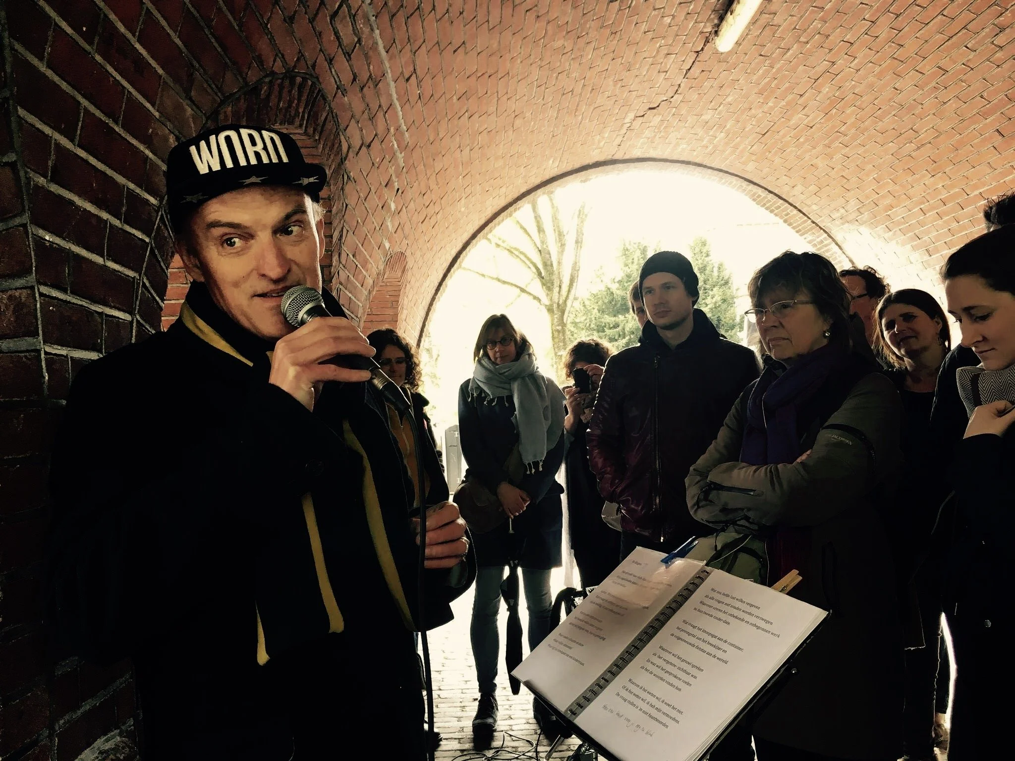 Een man met een pet die 'WORN' leest, houdt een microfoon vast en spreekt tot een groep mensen onder een boog van bakstenen. Een muziekblad ligt op een statief voor hem. Mensen luisteren aandachtig, sommigen maken foto's of video.