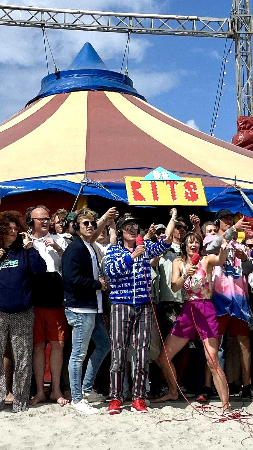 Een groep mensen staat samen voor een kleurrijke circustent met een bord dat 'CIRCUS' zegt, sommige mensen houden microfoons vast en dragen headphones, terwijl anderen plezier maken op de zandgrond onder een blauwe lucht.
