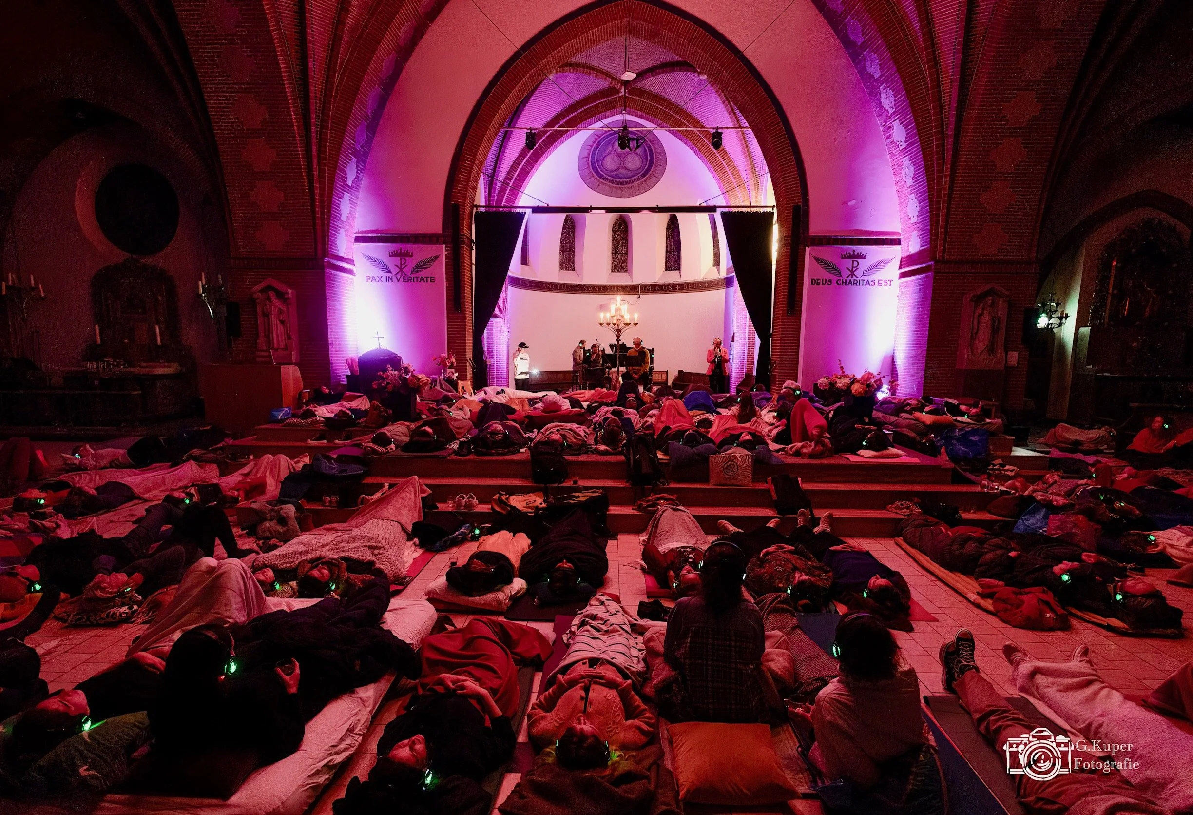 Een volle kerk met mensen die op de vloer liggen, waarschijnlijk in een gebeds- of meditatiekring, met mensen op het podium die iets presenteren of spreken. De kerk is verlicht met roze en paars licht, en er hangen banners met teksten in het Nederlands.