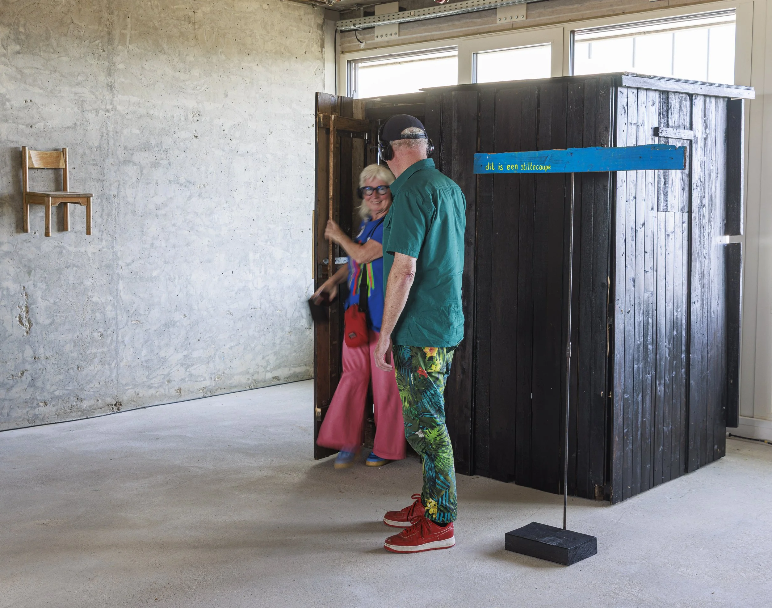 Twee mensen die een stiltecup aan het bekijken zijn in een ruimte onder bouw of renovatie, met een houten muur en een lege muur met een kleine houten stoel en een informele sfeer.
