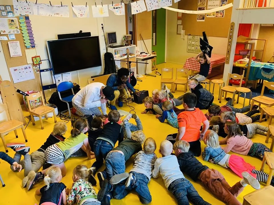 Klaslokaal met kinderen die op de vloer liggen luisteren naar twee mensen die iets presenteren met een microfoon. De kamer is kleurrijk met meubelen en decoraties.