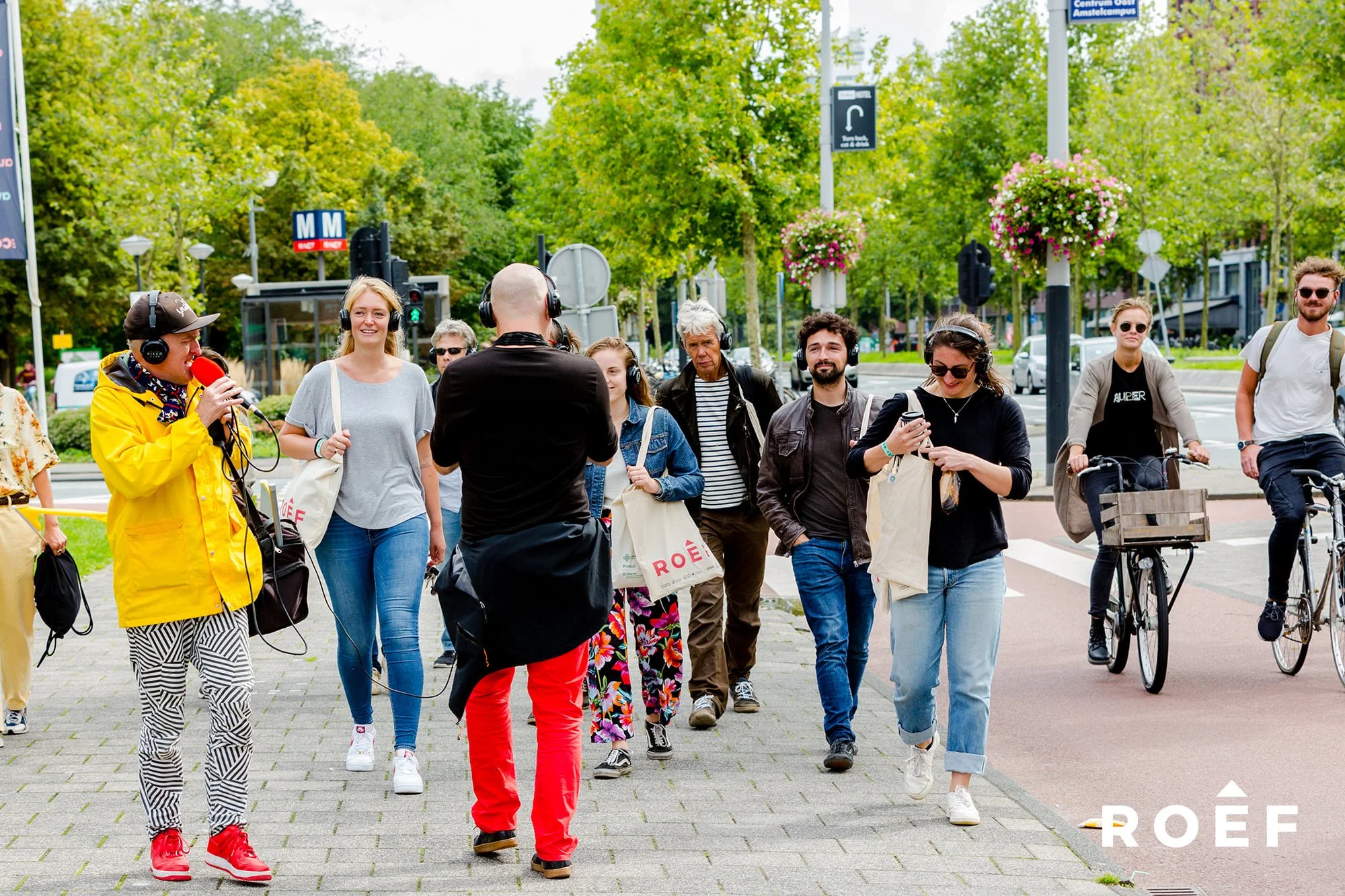 Groep mensen die een interview doen op straat tijdens een parkachtige omgeving met groene bomen en bloemen in hangmanden, met veel mensen die wandelen of fietsen op de achtergrond.