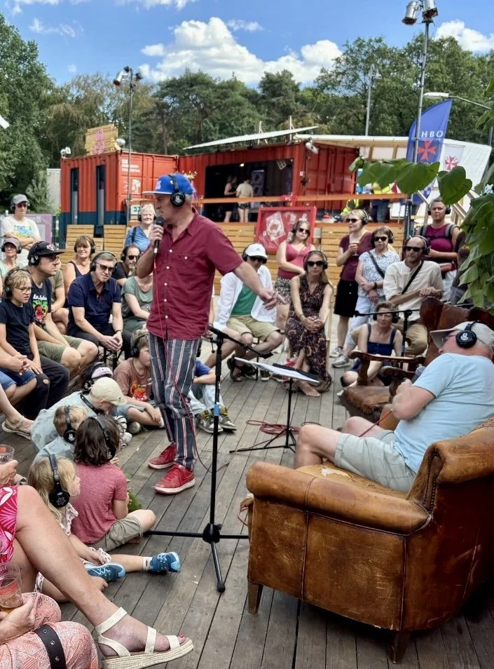 Man met blauwe hoed en koptelefoon zingt of spreekt in microfoon, omringd door een publiek op een houten terrasje, met een achtergrond van een rode container en amusementshuis.