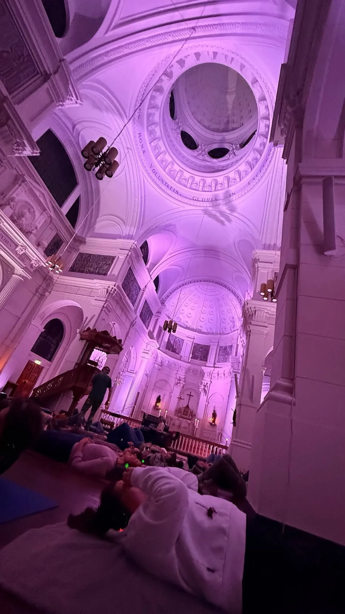 Binnenkant van een kerk of kathedraal met hoge koepels en gewelfde plafonds, verlicht in roze/paarse kleuren, met mensen die op de vloer rusten of bidden.
