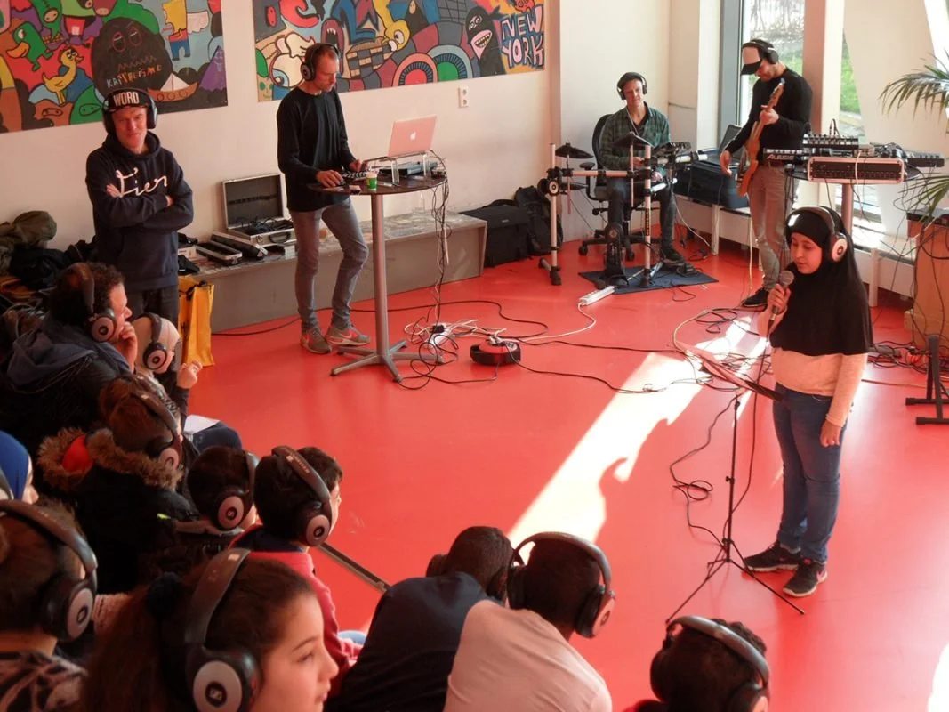 Een groep muzikanten en een zangeres met koptelefoons in een kamermuziek studio, met een publiek dat ook koptelefoons draagt.