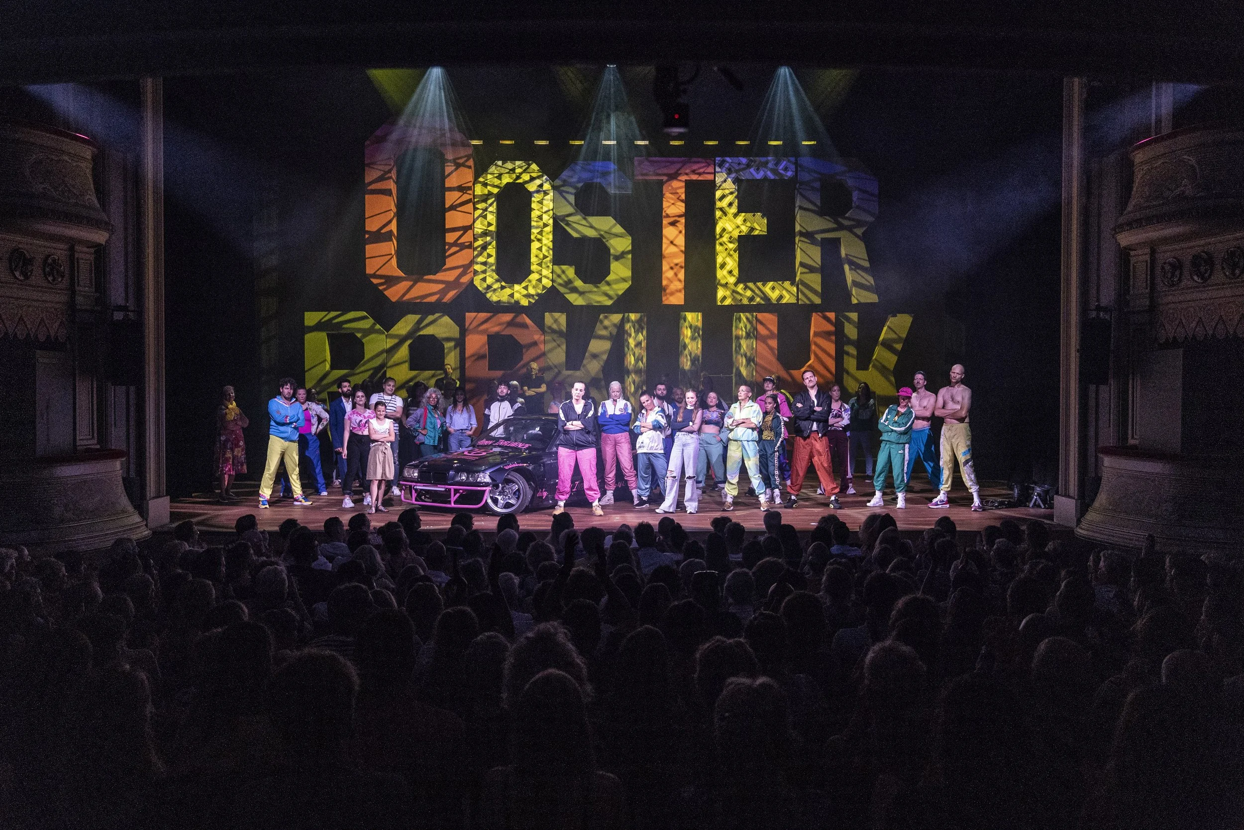 Een podium met een grote kleurrijke projectie die de woorden 'OOSTEN BEAT' toont, omringd door mannen en vrouwen in sportkleding en casual kleding, met een zwart sportauto vooraan.