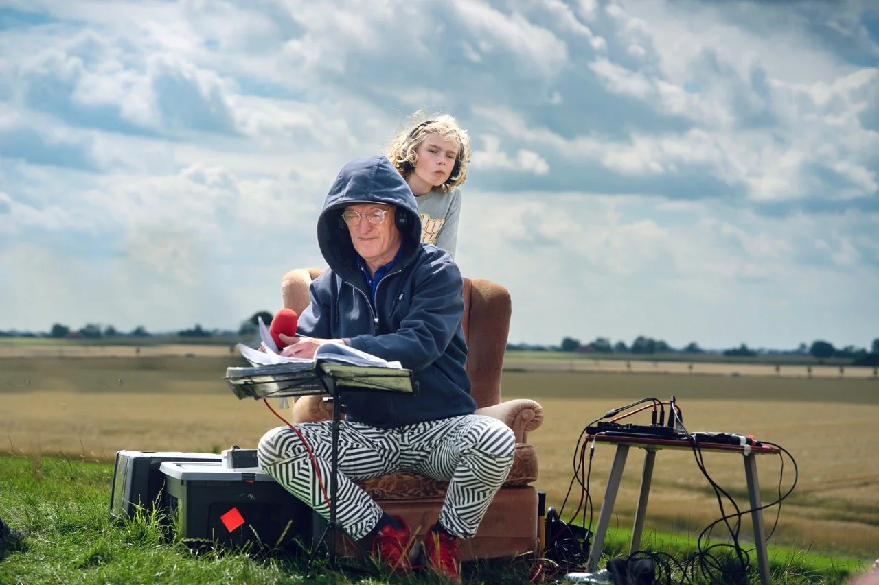 Man met koptelefoon en zonnebril zit op een stoel in een open veld, met een jong meisje op de schouder. Er bevindt zich apparatuur en papieren voor muzikale opnames.