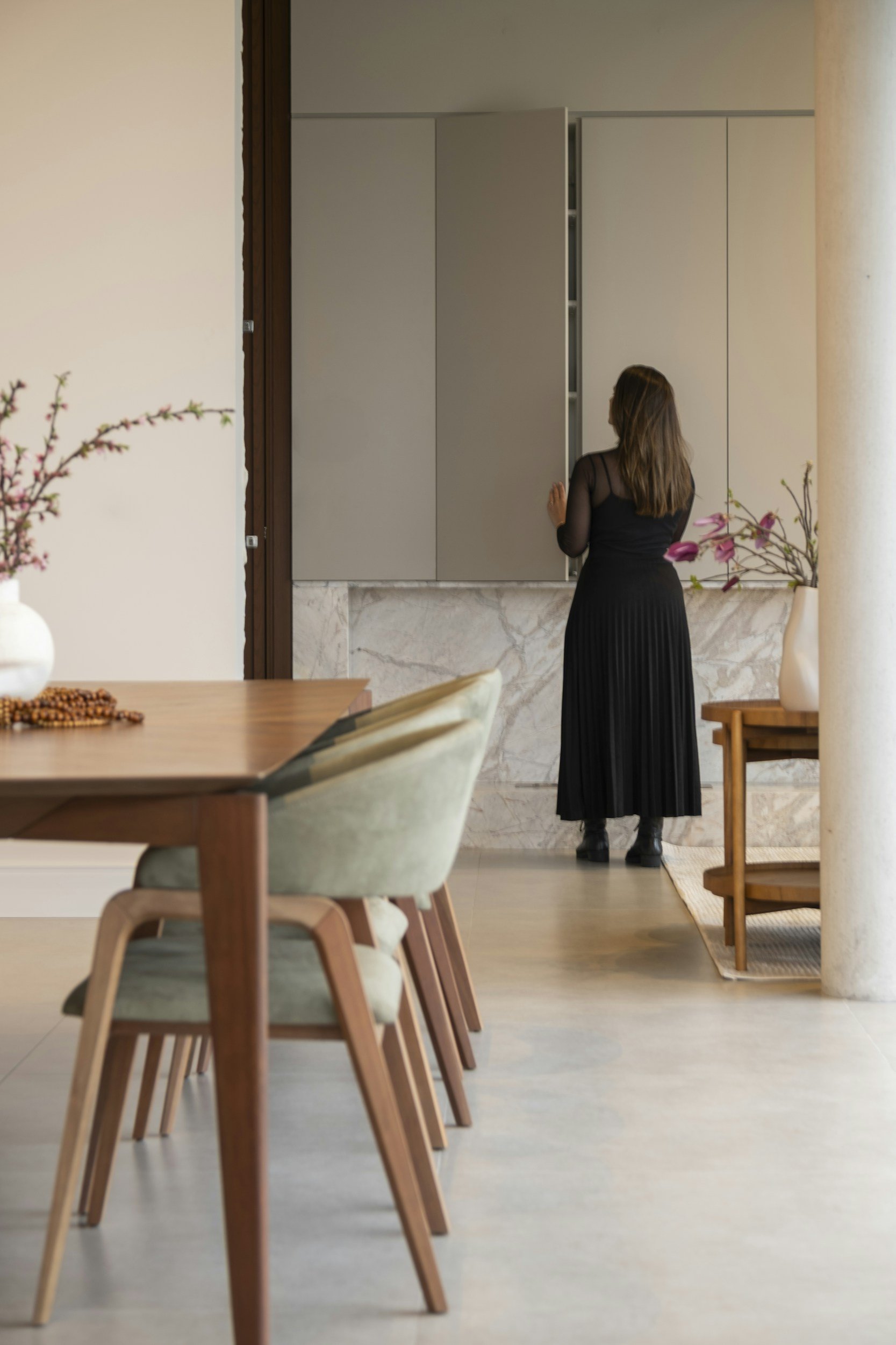 A woman in a black dress opening a tall cabinet in a kitchen, seen past a wooden dining table.