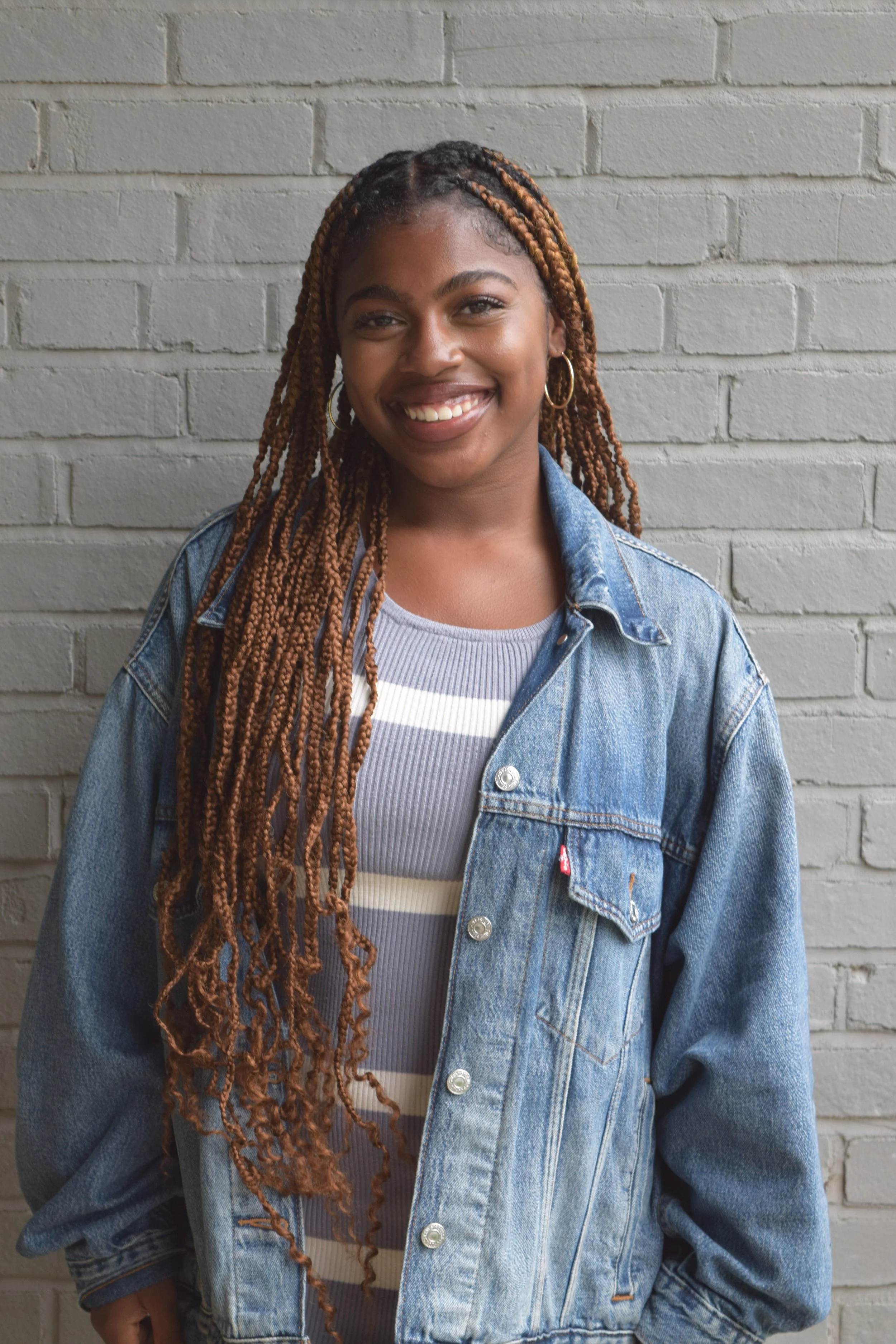 Morgan Hackett with long, braided hair, wearing a denim jacket and hoop earrings, smiling in front of a gray brick wall for her intership with Breakthrough Birmingham