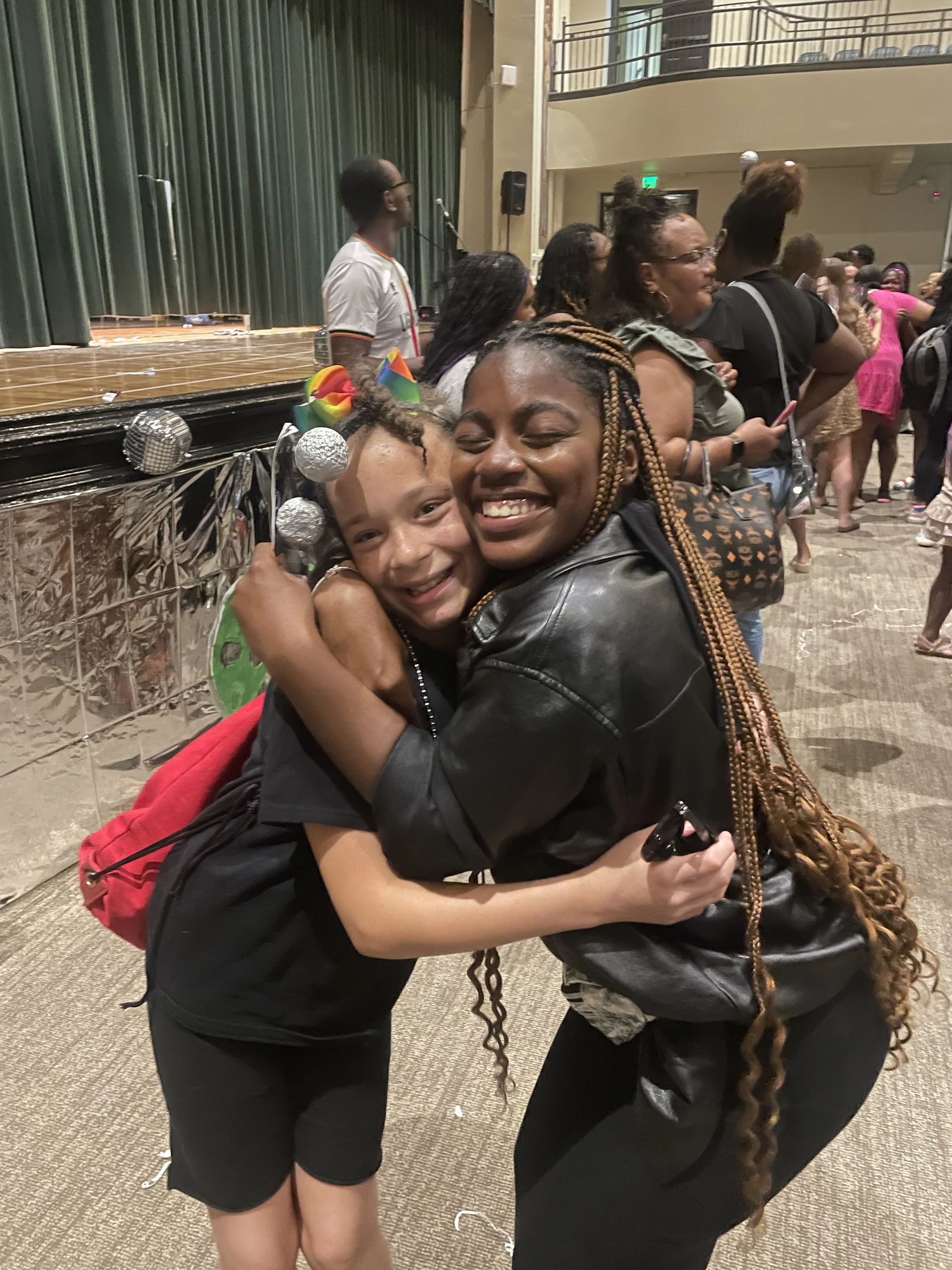 Two women hugging and smiling at a lively indoor event with a stage and people in line.
