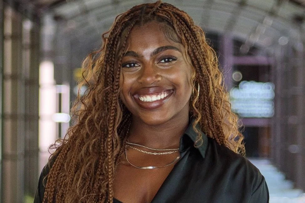 Portrait of a young woman with long, curly braids, smiling, wearing a black shirt, layered necklaces, standing in a subway station.