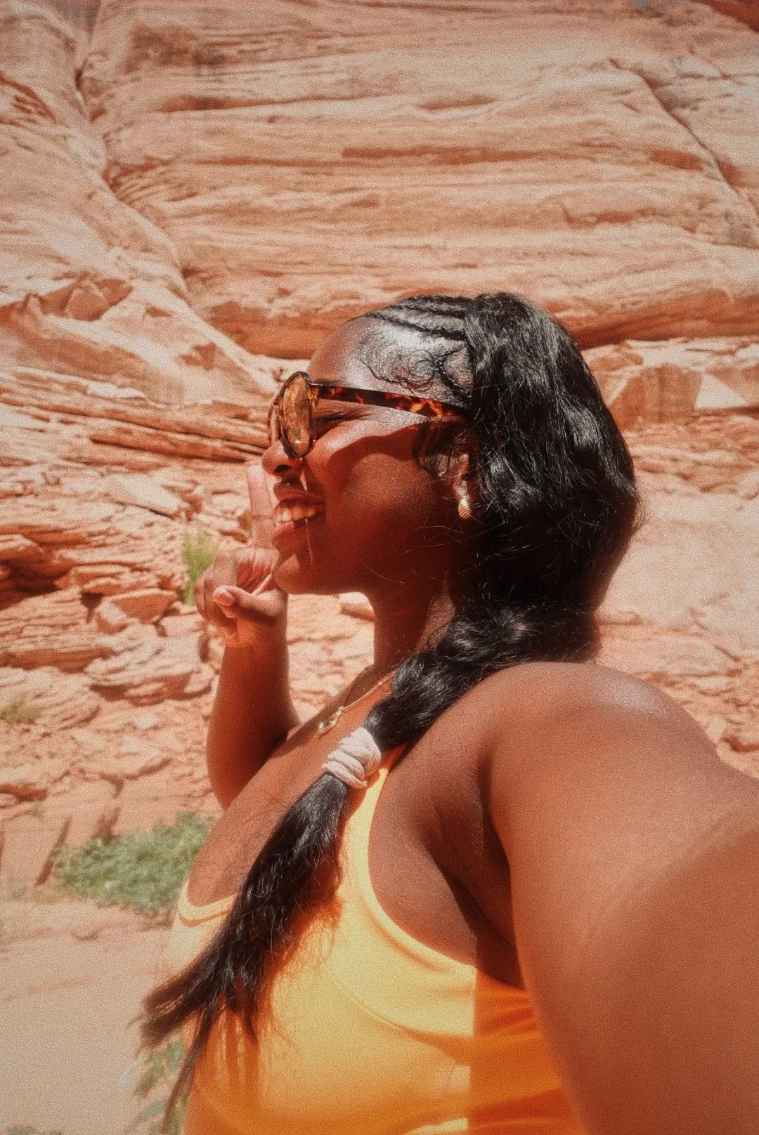 A woman with braided hair, wearing sunglasses and a yellow tank top, smiling and making a shushing gesture with her finger against a desert red rock landscape background.